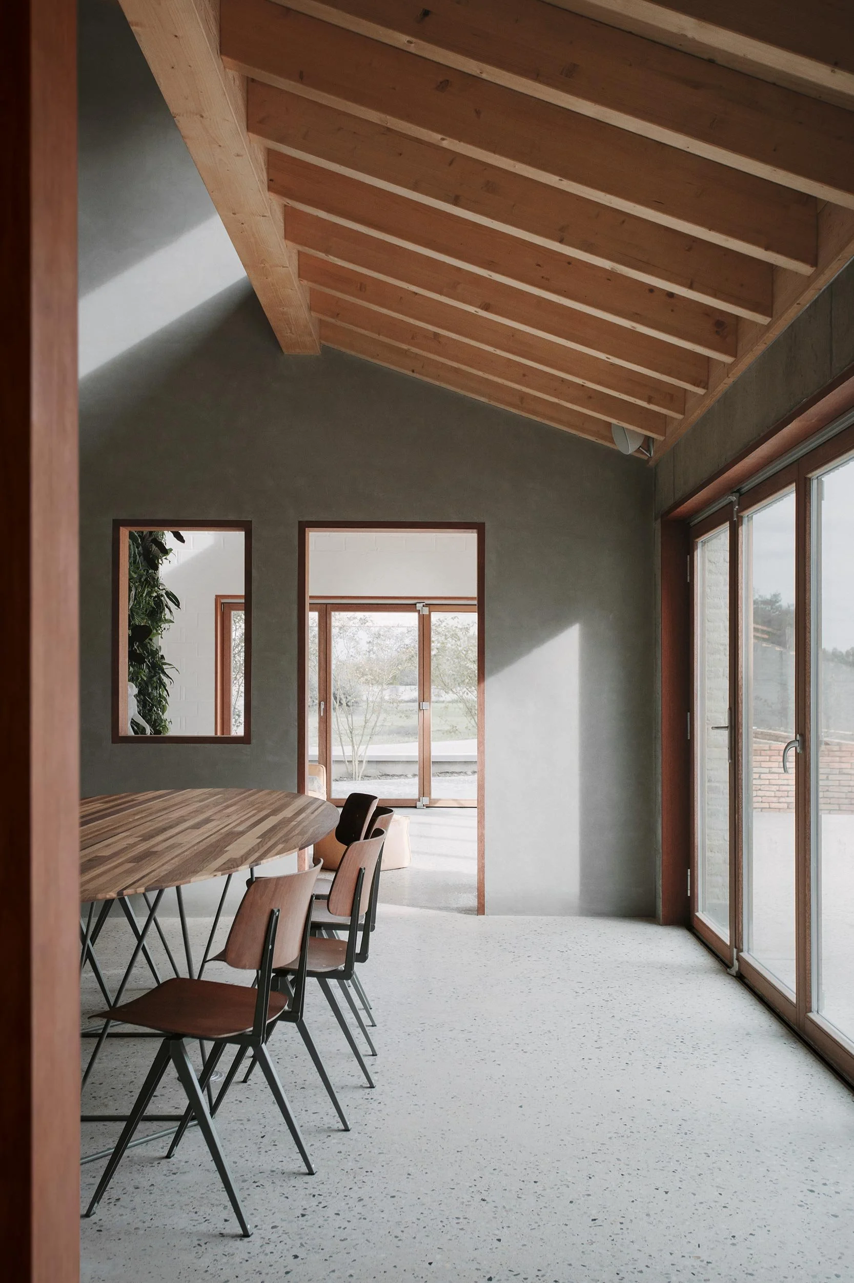 Contemporary dining room in a renovated farmhouse featuring an exposed wooden ceiling, terrazzo flooring, and large glazed openings that bring natural light onto handmade wooden furniture.