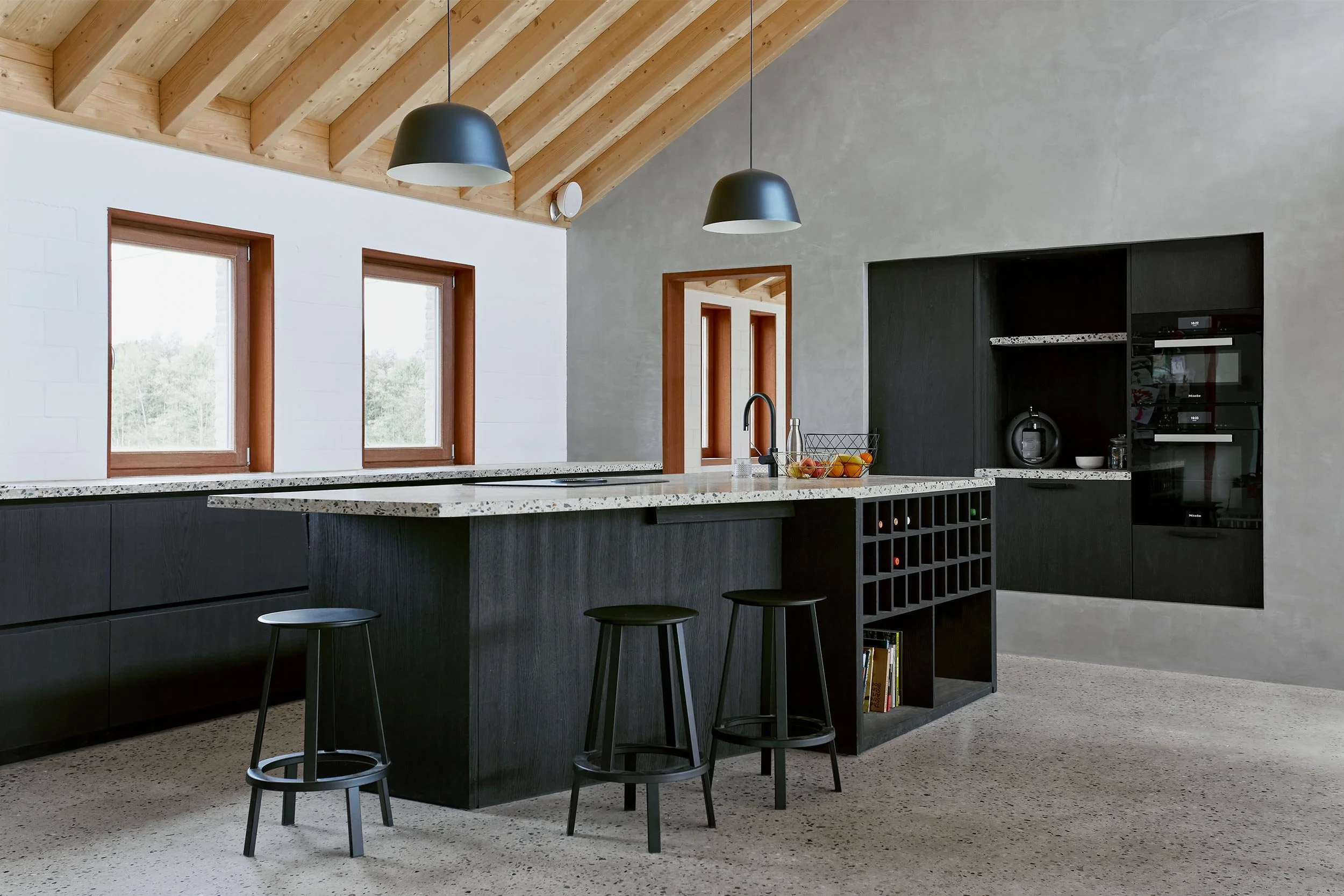 Contemporary kitchen inside a renovated farmhouse with exposed wooden beams, terrazzo countertop, dark cabinetry, and natural light.