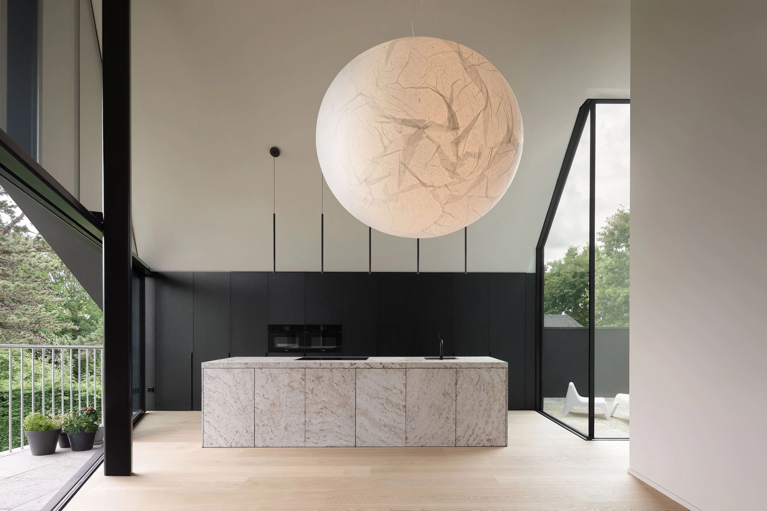 modern kitchen with marble island, black wooden cabinets, and a spherical Moon lamp by Davide Groppi, opening to a terrace with white chairs, plants, and a large panoramic window overlooking nature