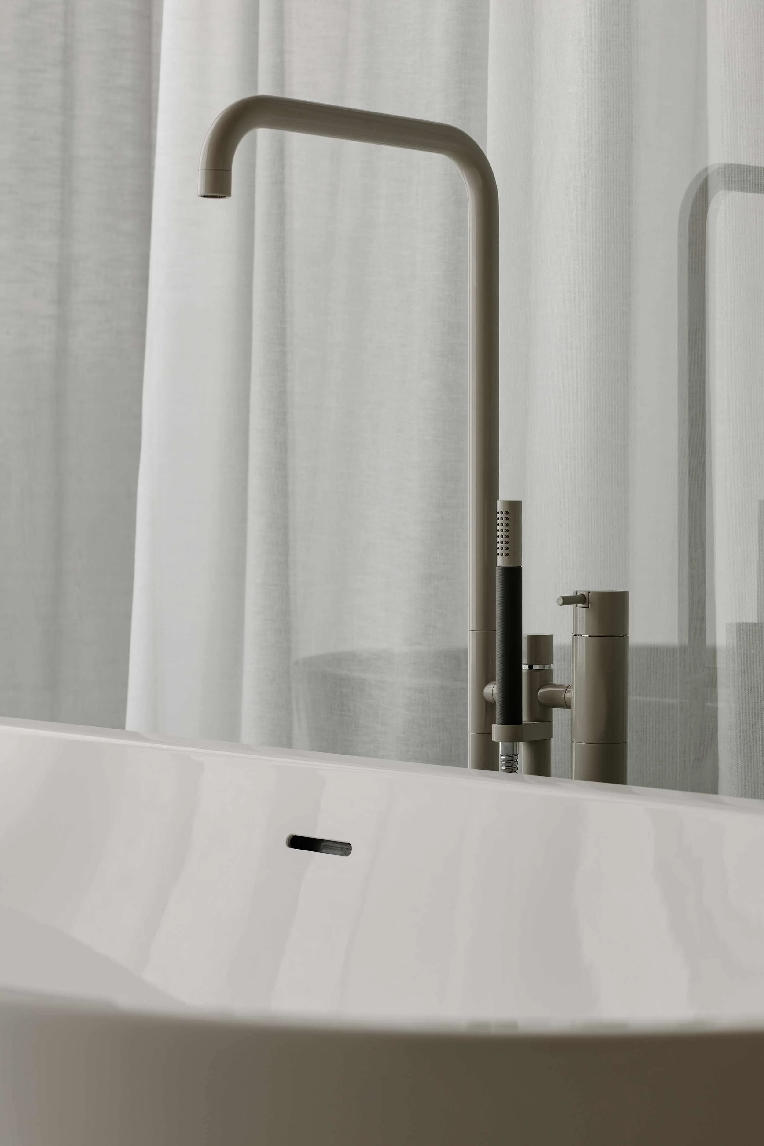Minimalist bathroom with a white freestanding Vola bathtub and sculptural brushed metal bath tap against white curtains.