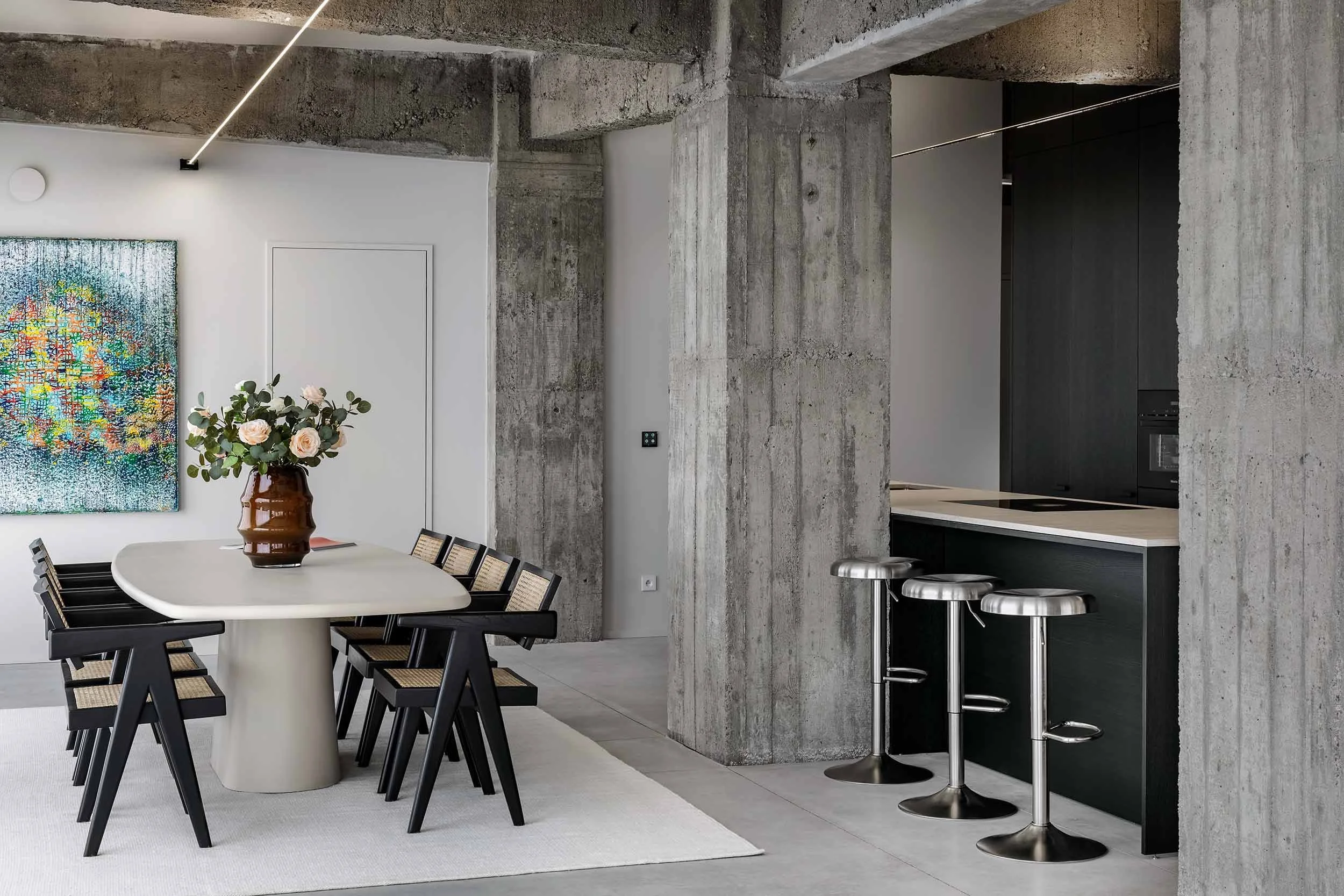 Modern kitchen with concrete walls behind a dining area, featuring a round white table with a brown vase of pink and white flowers, and black dining chairs. The kitchen has a dark counter with bar stools and sleek black cabinets, with a colorful abstract artwork on a white wall.
