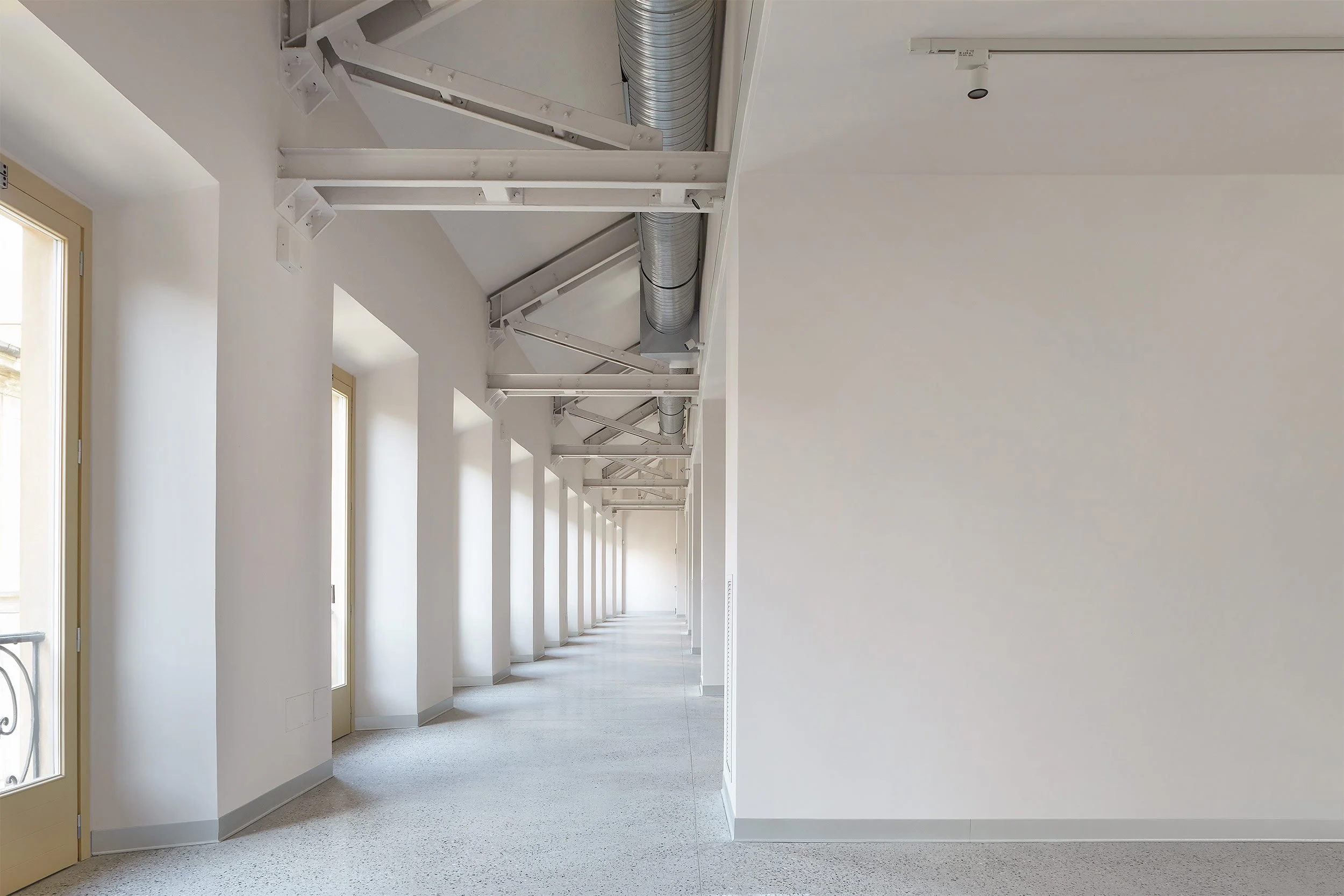 Corridoio restaurato di Palazzo Citterio a Milano, caratterizzato da pareti bianche, infissi chiari, luce naturale proveniente dalle grandi finestre e soffitto con travi metalliche a vista e impianti esposti.