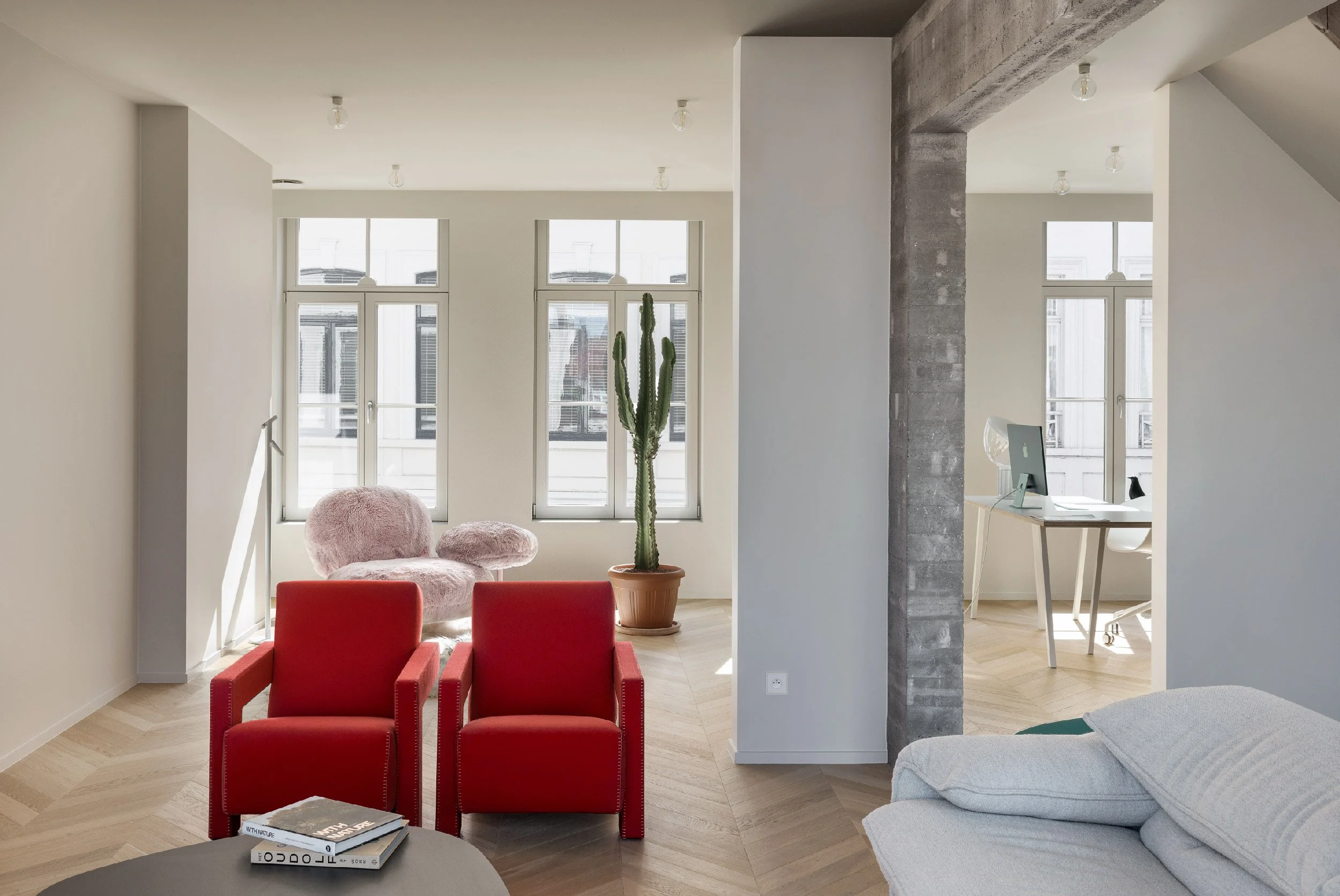 Light-filled interior of renovated house in Bruges with white walls, light wood parquet floors, beige Cassina Maralunga sofa, red Utrecht armchairs by Gerrit Thomas Rietveld, Edra Cipria chair, Flos Taccia lamp, and a potted cactus.