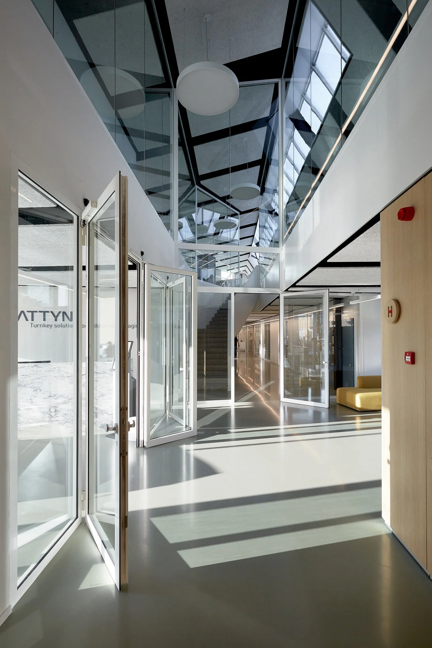 Interior view of Pattyn headquarters featuring a modern office space with extensive glass structures, concrete flooring, open staircases, and natural light creating a bright and transparent architectural environment.