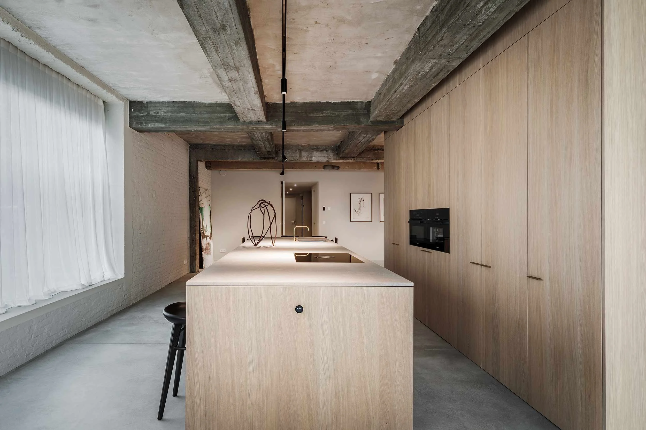 Modern kitchen with a large light wood island, black stool, and a built-in oven, featuring an exposed concrete ceiling and wooden cabinetry.