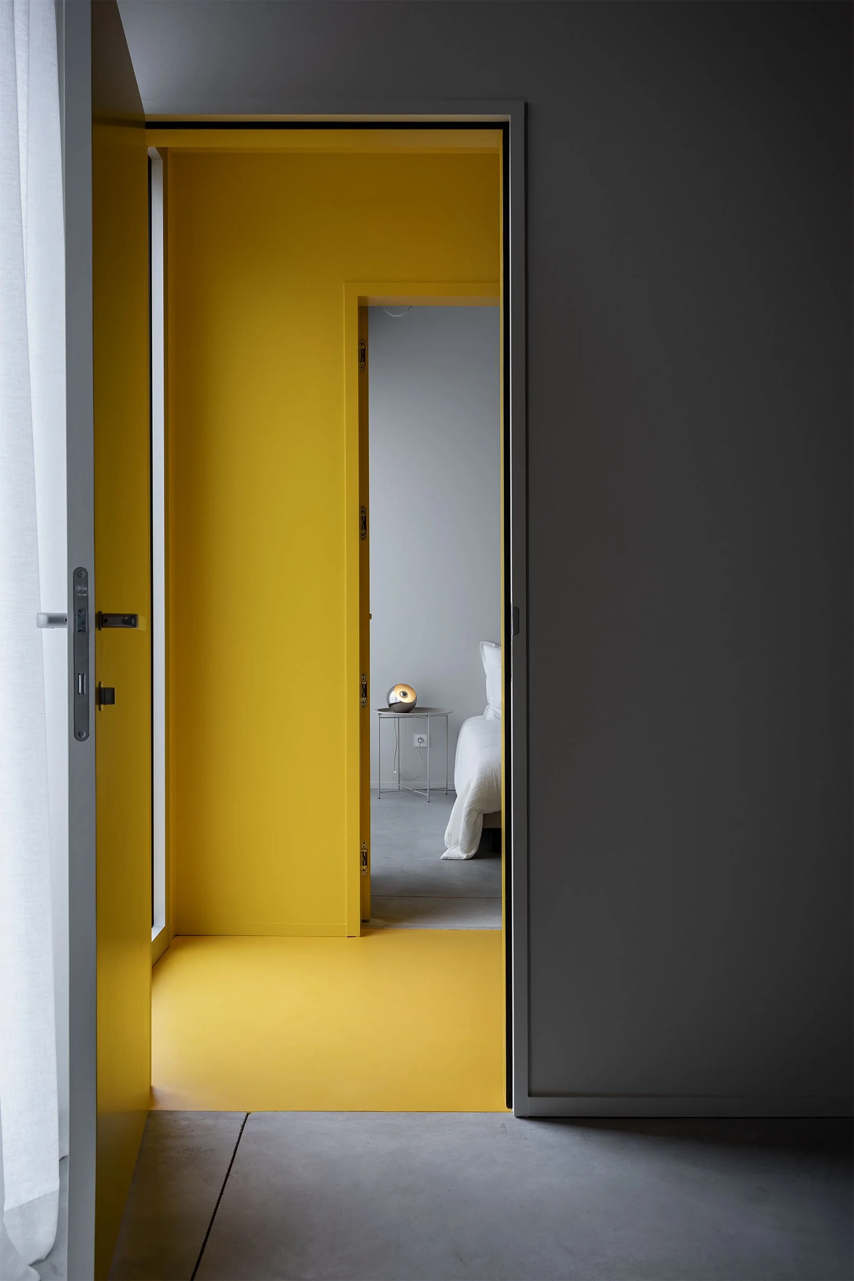 Zicht vanuit de gang op een badkamer met gele wanden en een slaapkamer op de achtergrond, met ruwe betonnen kolommen in een industrieel interieur.