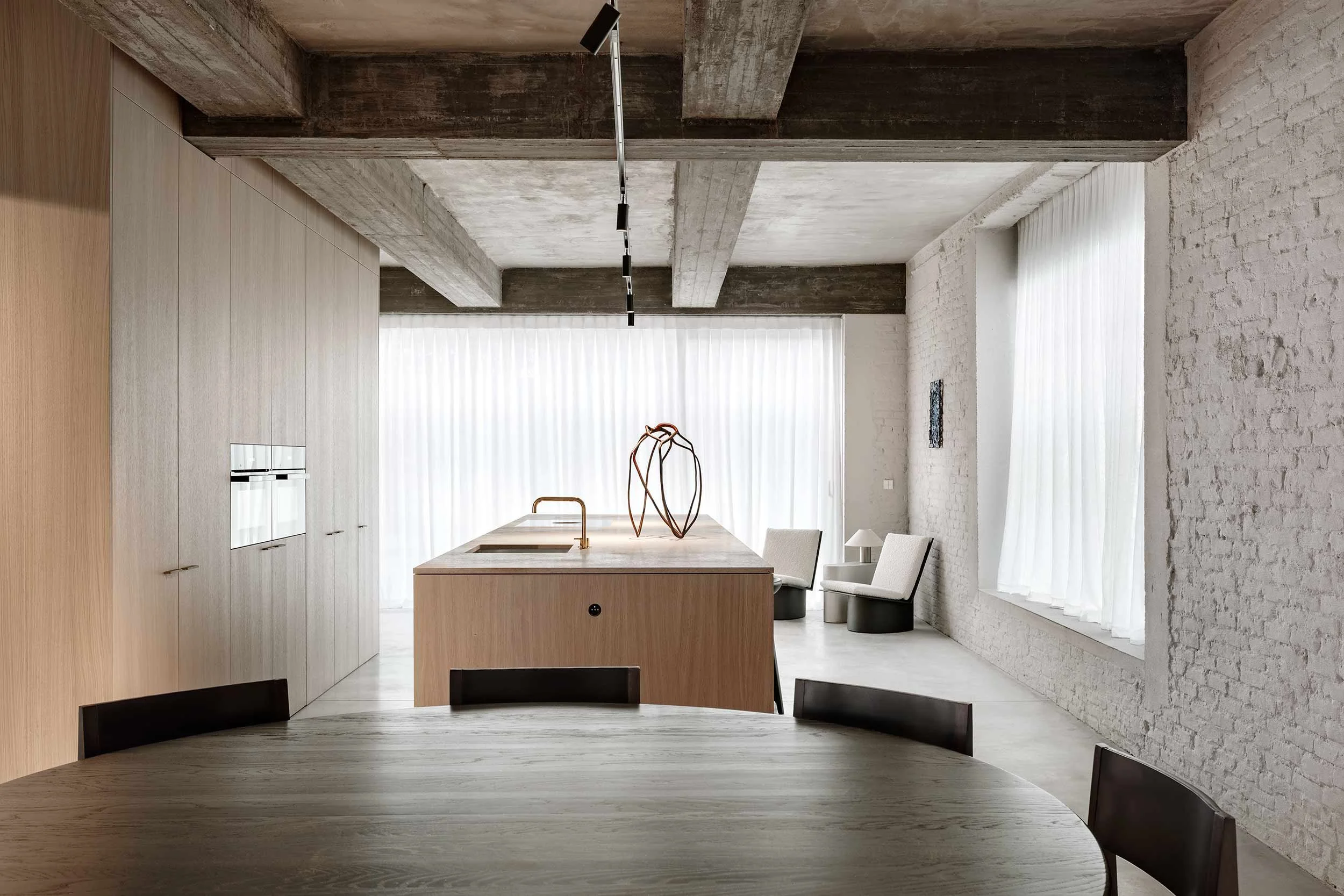 Modern kitchen with a large island, light wood cabinets, and white brick walls, illuminated by natural light through large windows with sheer curtains.