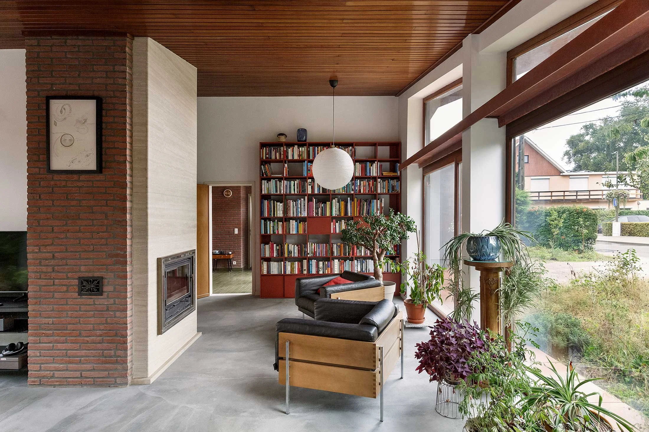 Living room with a smooth béton concrete floor, large windows overlooking the garden, and a brick fireplace, blending 1970s architecture with modern design.