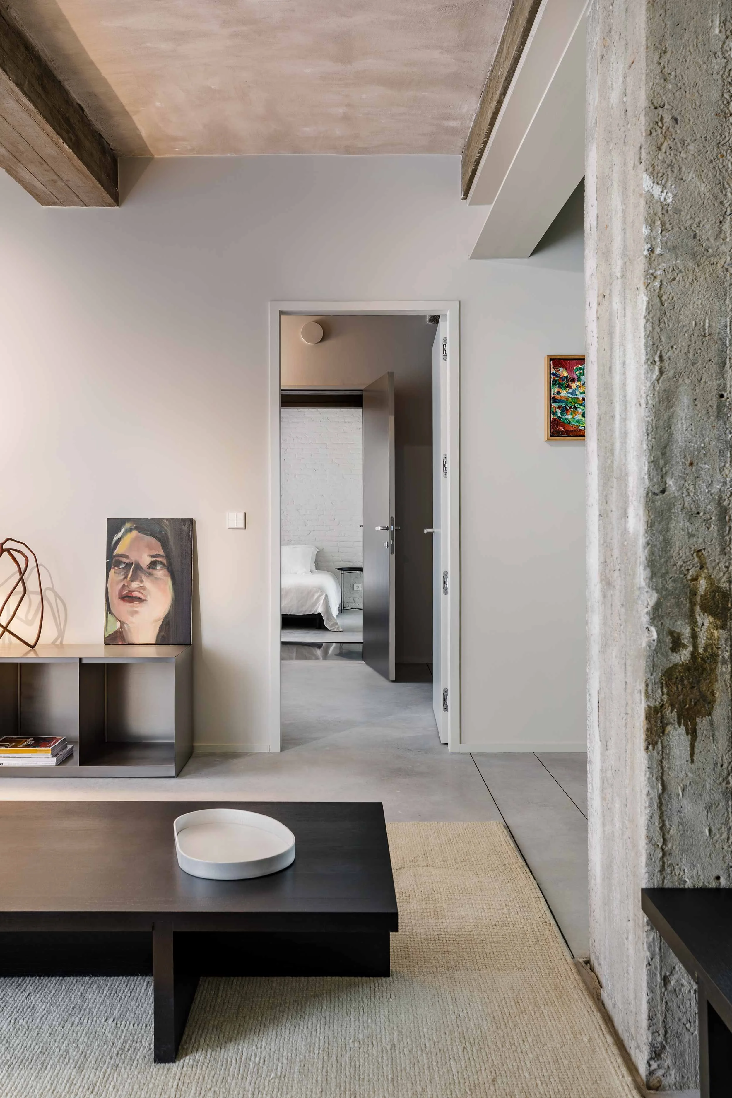 Frontaal zicht op een modern interieur met een betonnen kolom, een salontafel op de voorgrond en een gedeeltelijk zicht op de woonkamer en de slaapkamer op de achtergrond.