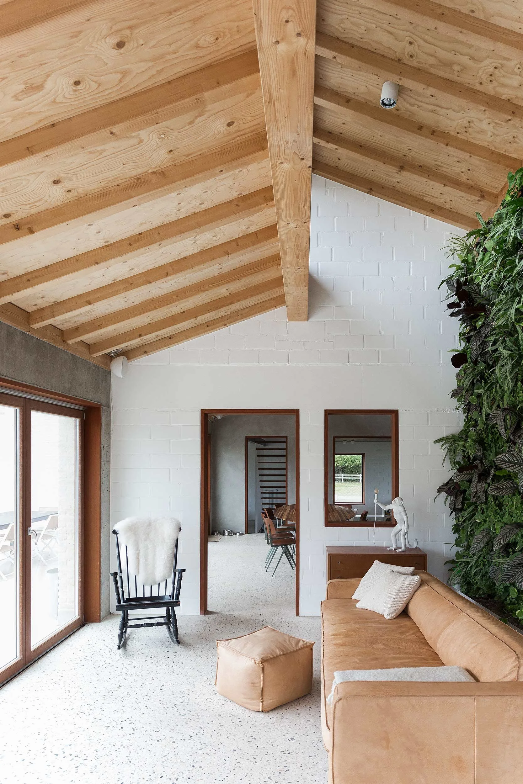 Contemporary living room in a renovated farmhouse featuring exposed wooden ceiling beams, terrazzo flooring, natural light, and a warm interior with leather sofa, indoor green wall, and minimalist wooden furnishings.