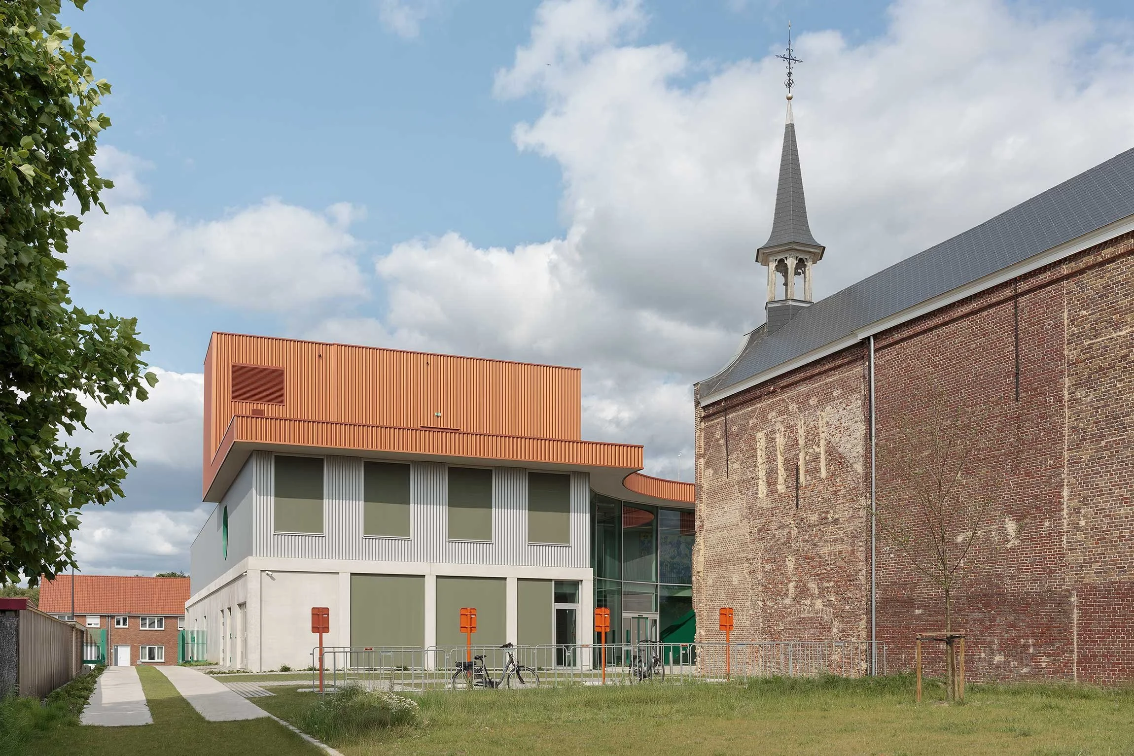 Modern building with white walls and large windows, clad in orange metal, next to an ancient brick wall. The contrast between the sleek orange and gray facade and the historic chapel reinforces the site’s unique architectural identity.