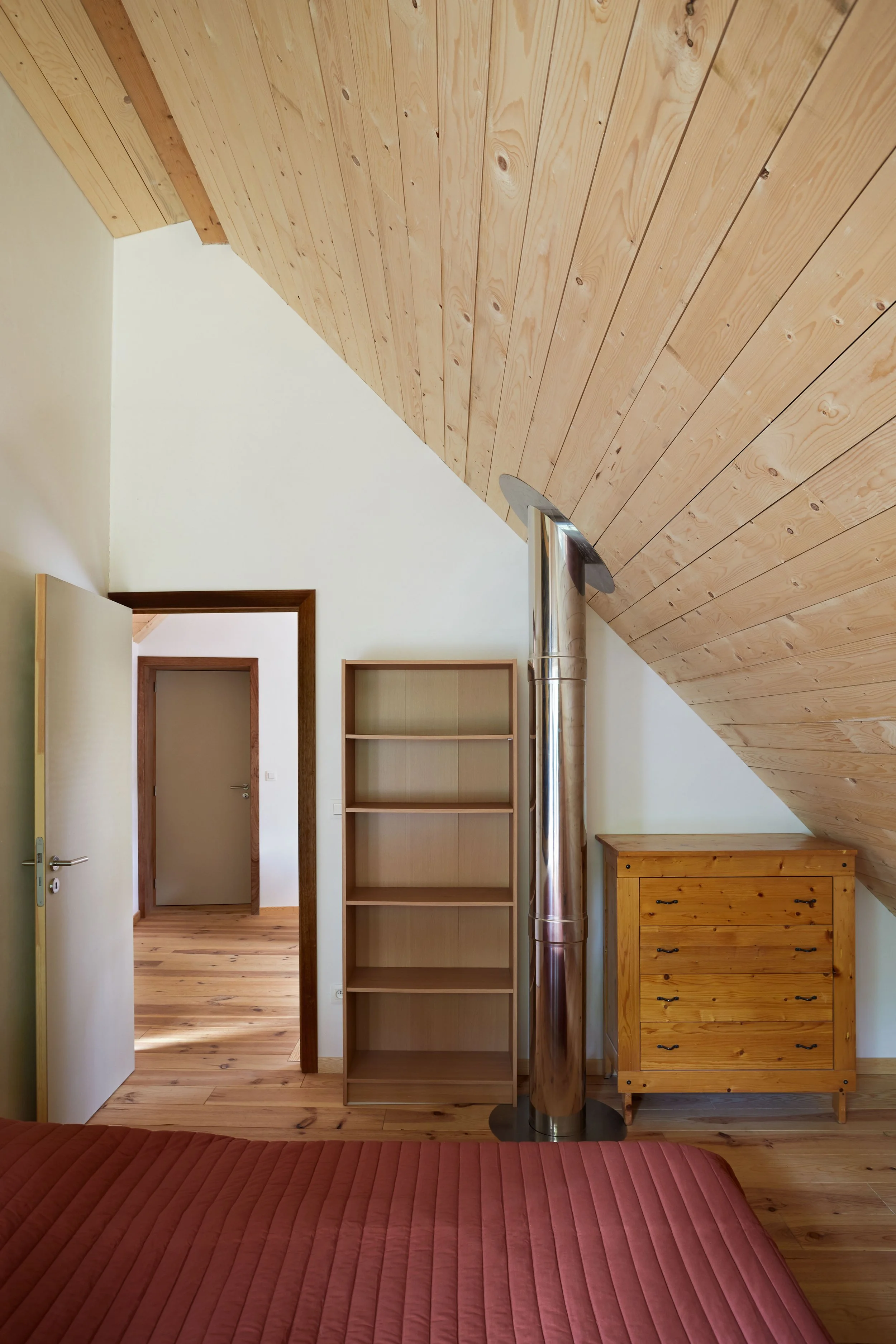Bedroom of an eco-friendly wooden house in the forest, featuring a sloped wooden ceiling, stainless steel chimney, wooden furniture, and a red quilt, with an open door leading to a wood-lined corridor.