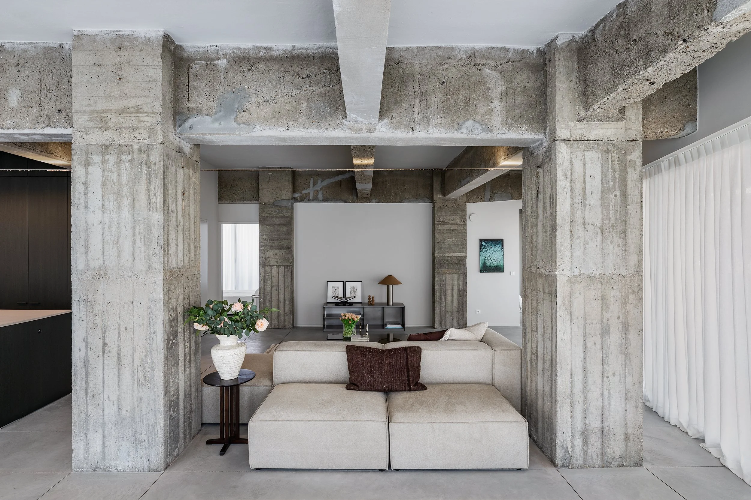 Living room with concrete beams and columns, beige sectional sofa, white vase with flowers, side table, wall art, and white curtains.