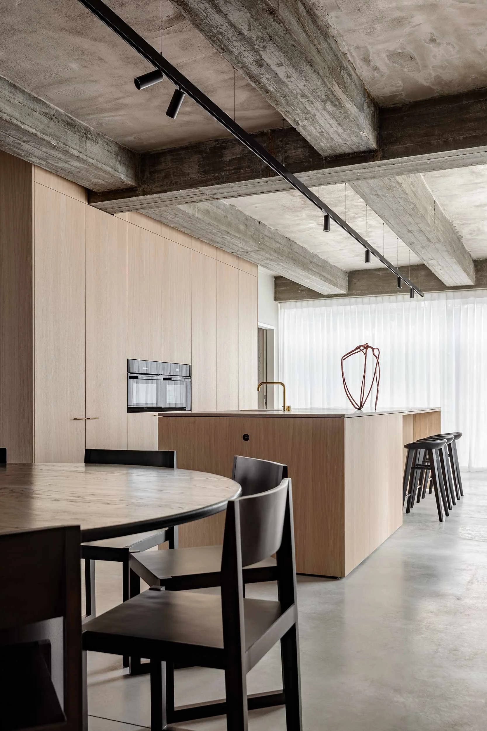 Modern kitchen with concrete ceiling and light wood cabinetry. Black chairs around a light wood table, and a kitchen island with bar stools. Minimalist decor and natural light.