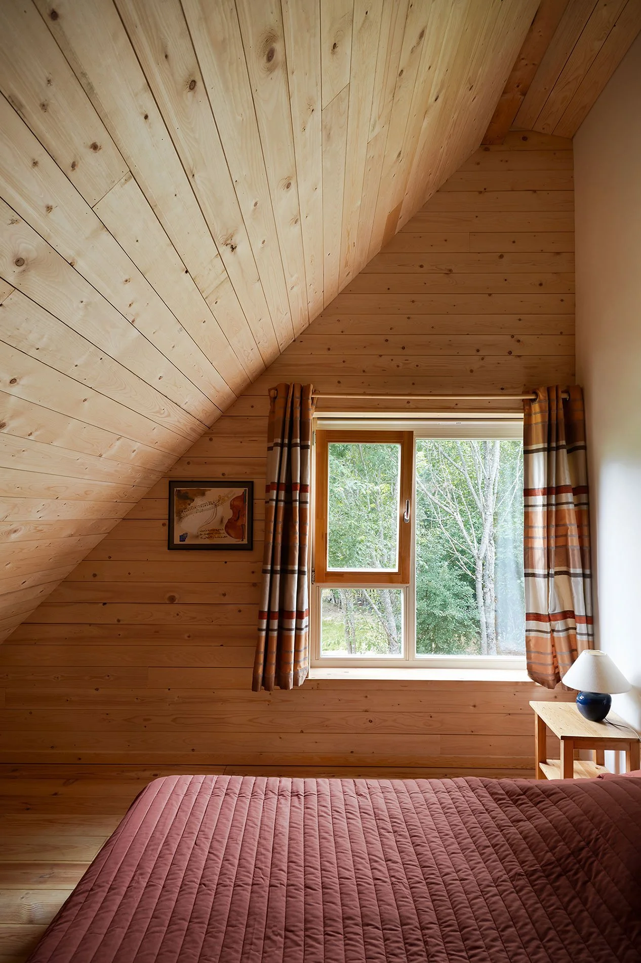 Sustainable forest cabin bedroom with sloped wooden ceiling, natural wood walls, a large window overlooking the trees, warm curtains, and a red quilt on the bed.