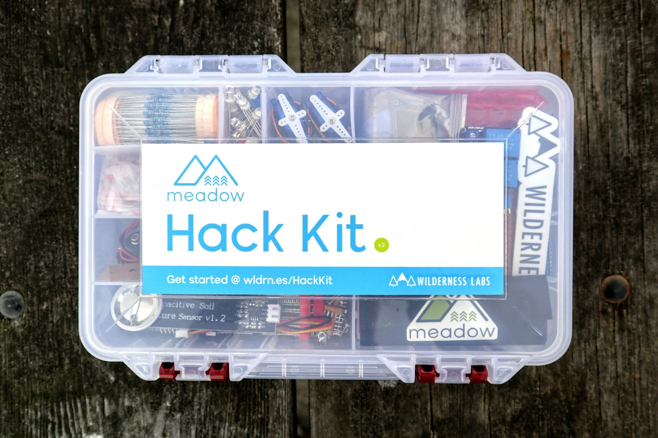 A clear plastic container labeled 'Hack Kit' from Wilderness Labs, placed on a weathered wooden surface. The kit contains electronic components, wires, and tools for DIY projects.