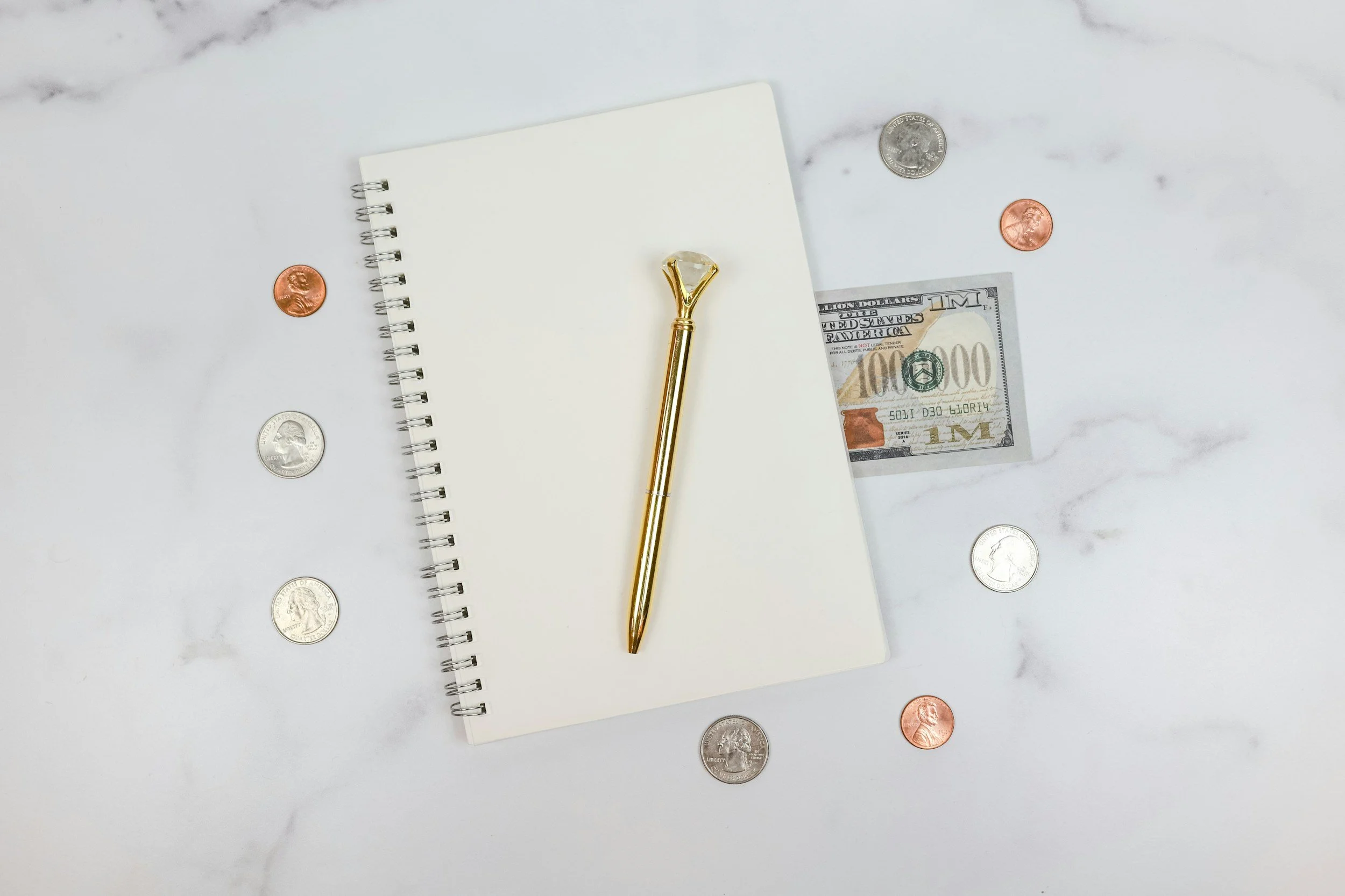 A white marble surface with a spiral notebook, a gold and diamond roller, U.S. dollar bills, and scattered U.S. coins.