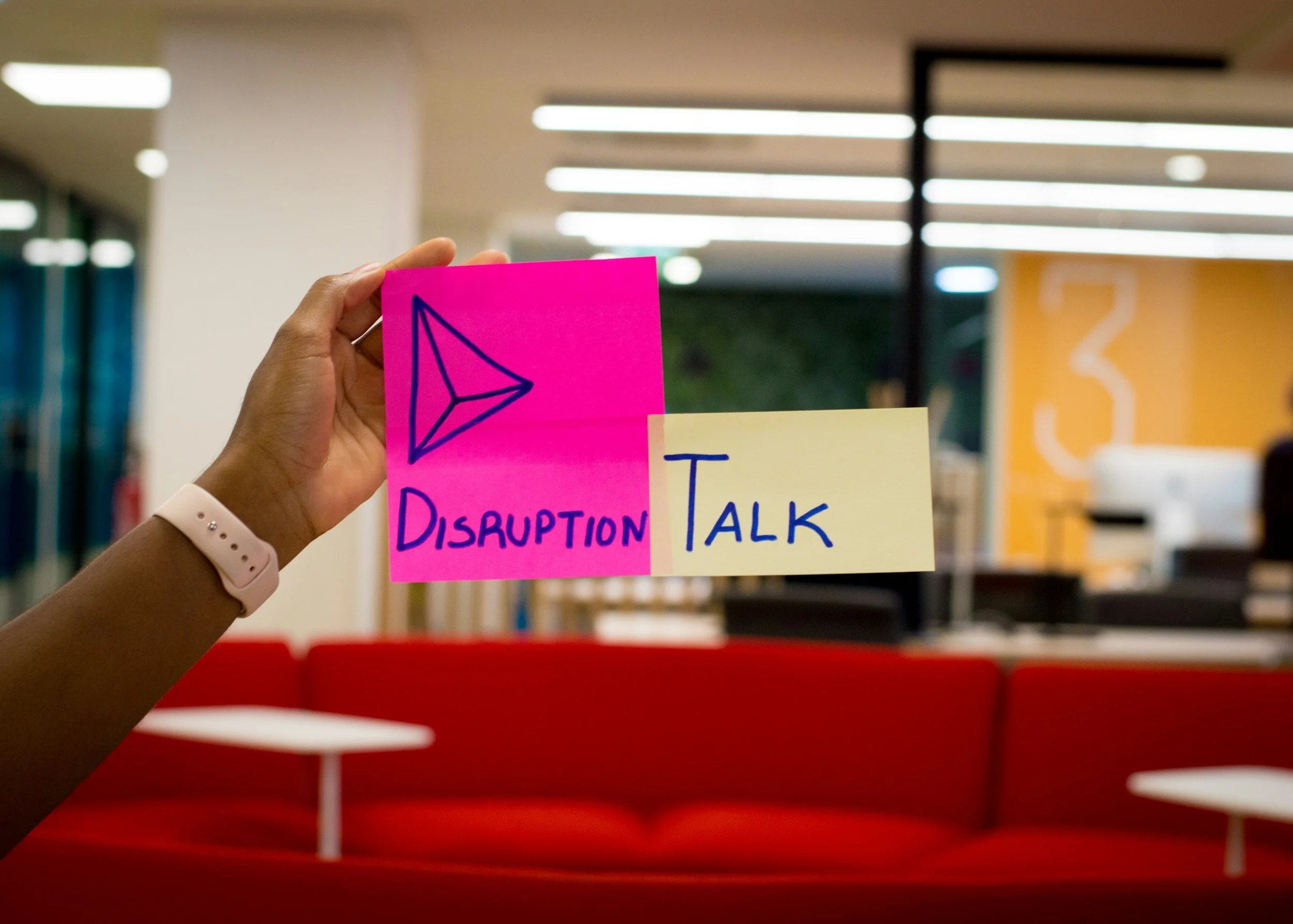 A person holding a bright pink sticky note with a blue arrow drawing and the word 'Disruption' written underneath it, in a modern indoor setting with ceiling lights and colorful walls.