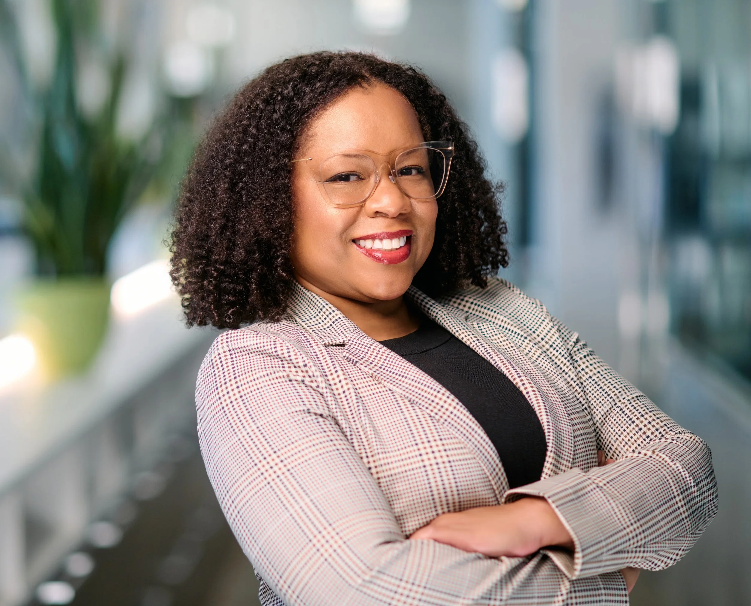 A smiling woman with curly dark hair, wearing glasses and a blazer, sitting with her arms crossed in an office environment with blurred background.