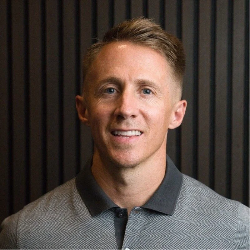 Headshot of a clean shaven man and medium length hair, smiling and wearing a grey collared shirt against a dark, textured background.