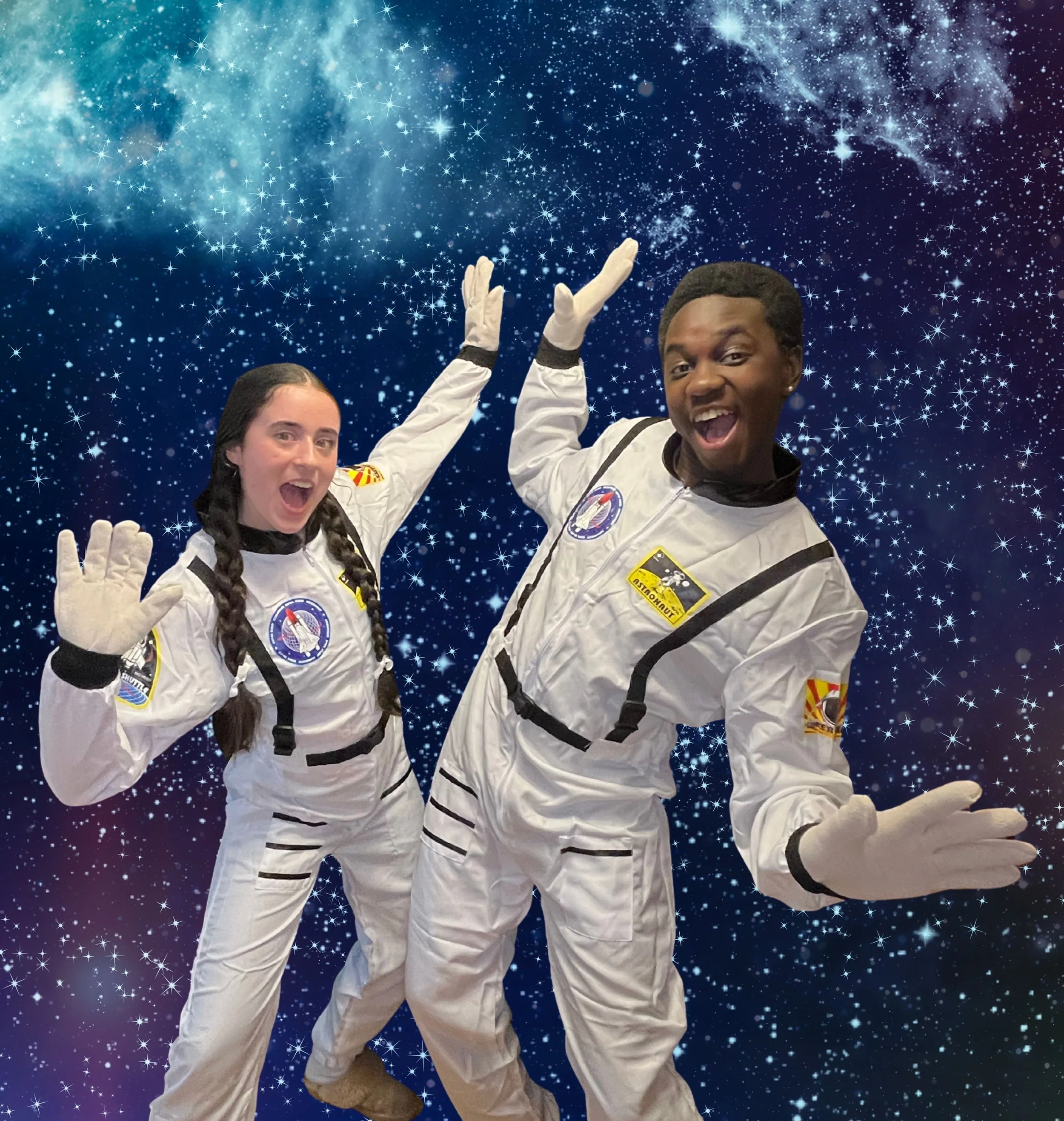 Two people dressed as astronauts smiling and posing in front of a starry galaxy backdrop with their hands raised.