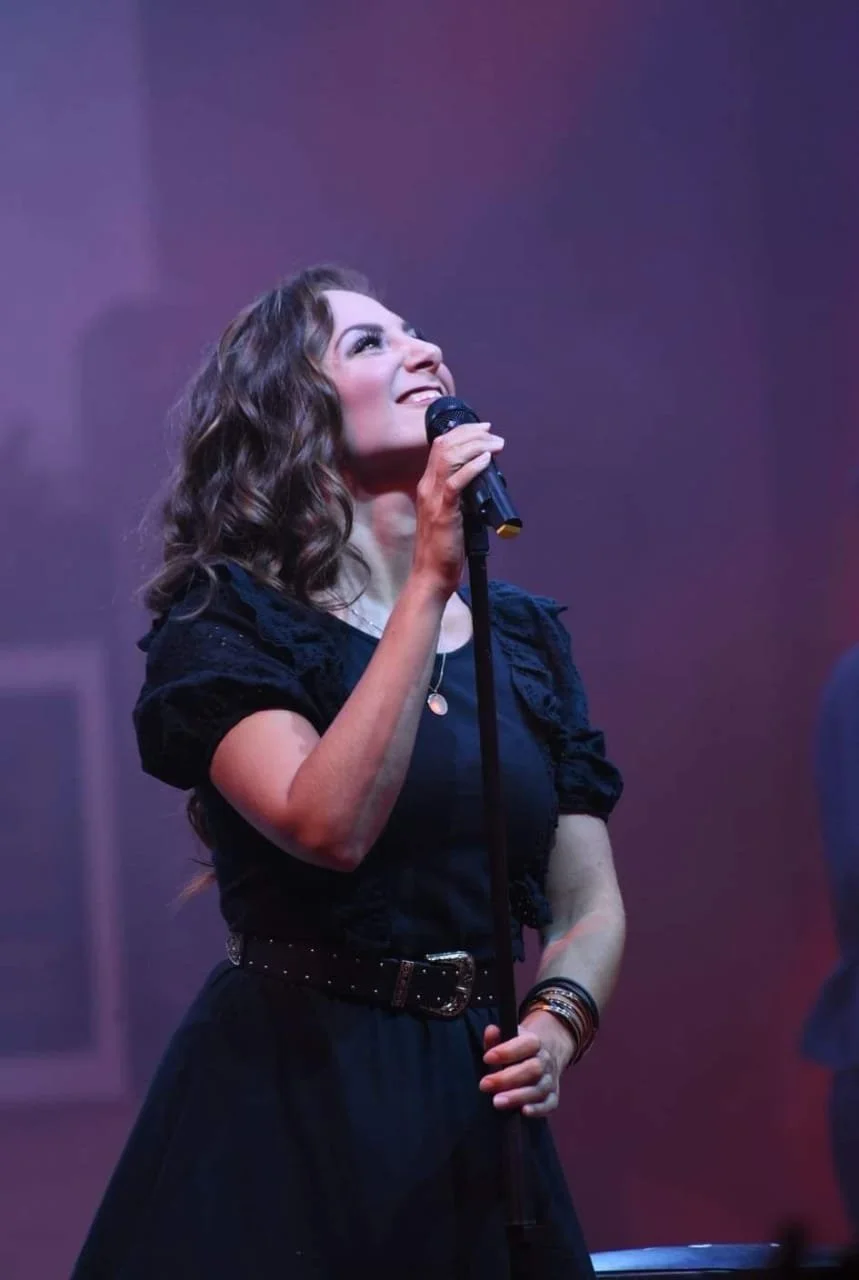 A woman singing into a microphone on stage, with a smile and a joyful expression, in a dark-colored outfit with ruffled sleeves and a belt, against a purple background.