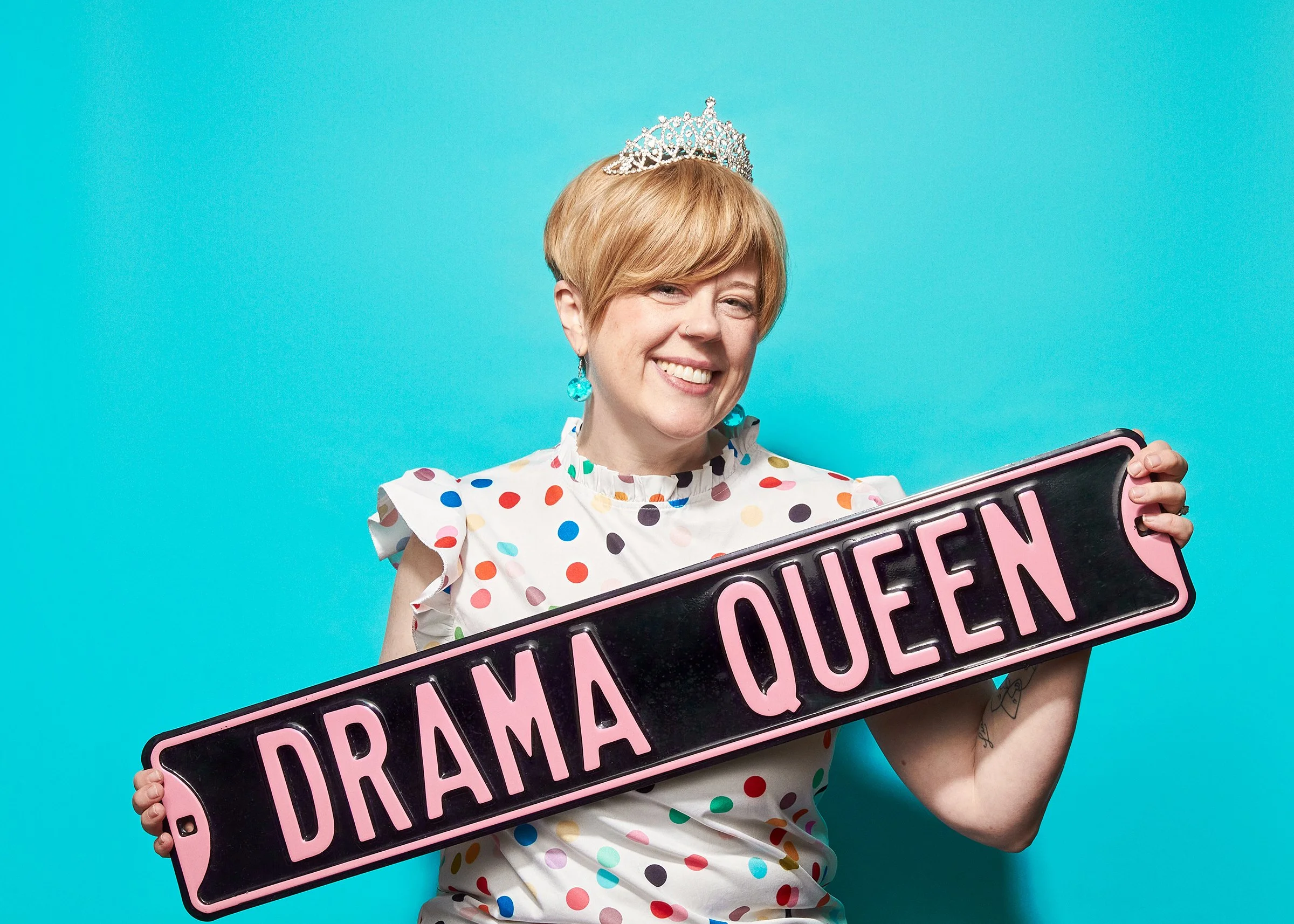 A woman with short blonde hair wearing a tiara and a white dress with colorful polka dots, smiling and holding a sign that reads 'Drama Queen' against a bright blue background.
