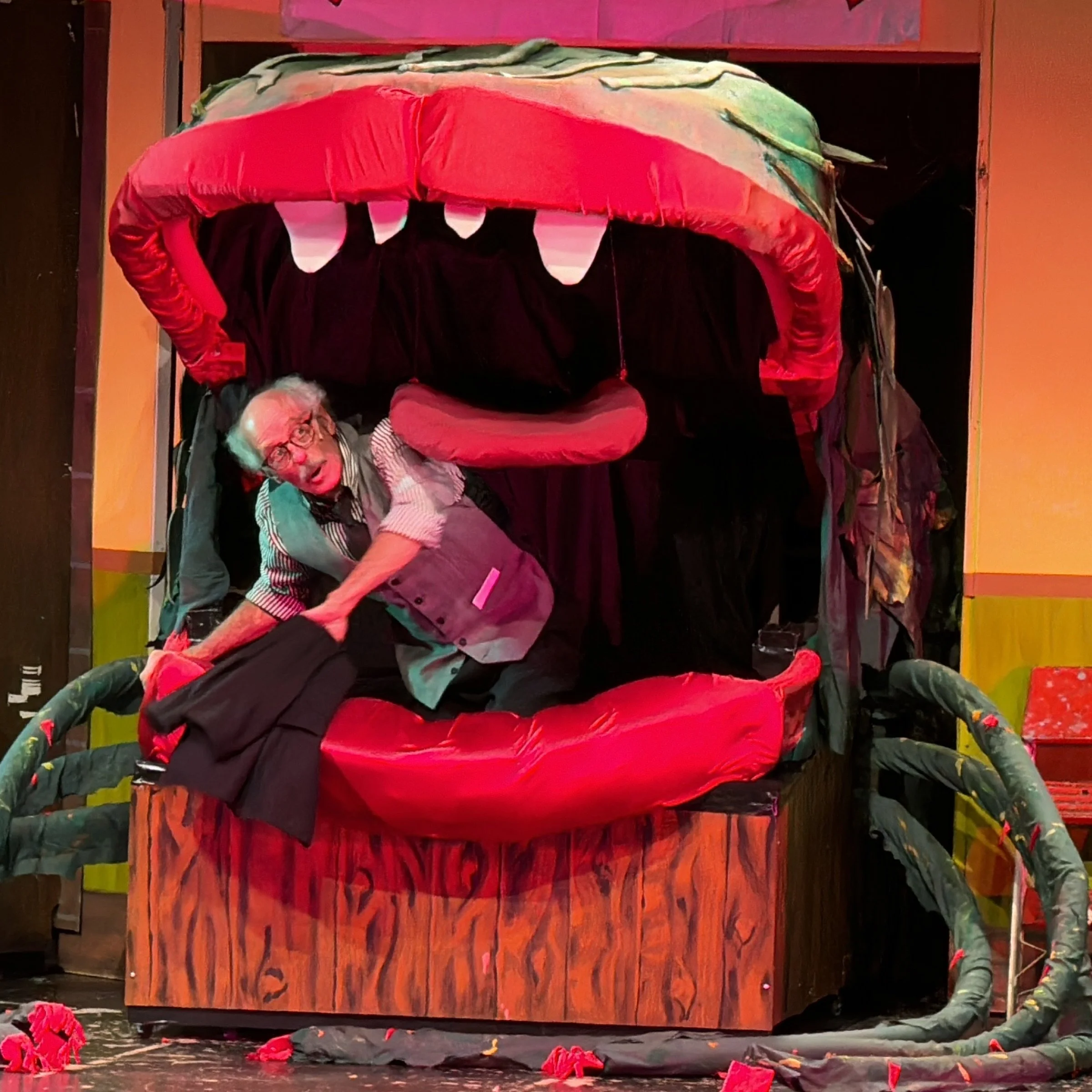 A man with glasses, gray hair, and a striped shirt, appears to be inside a large puppet or costume resembling an open mouth with sharp teeth, part of a theatrical set.