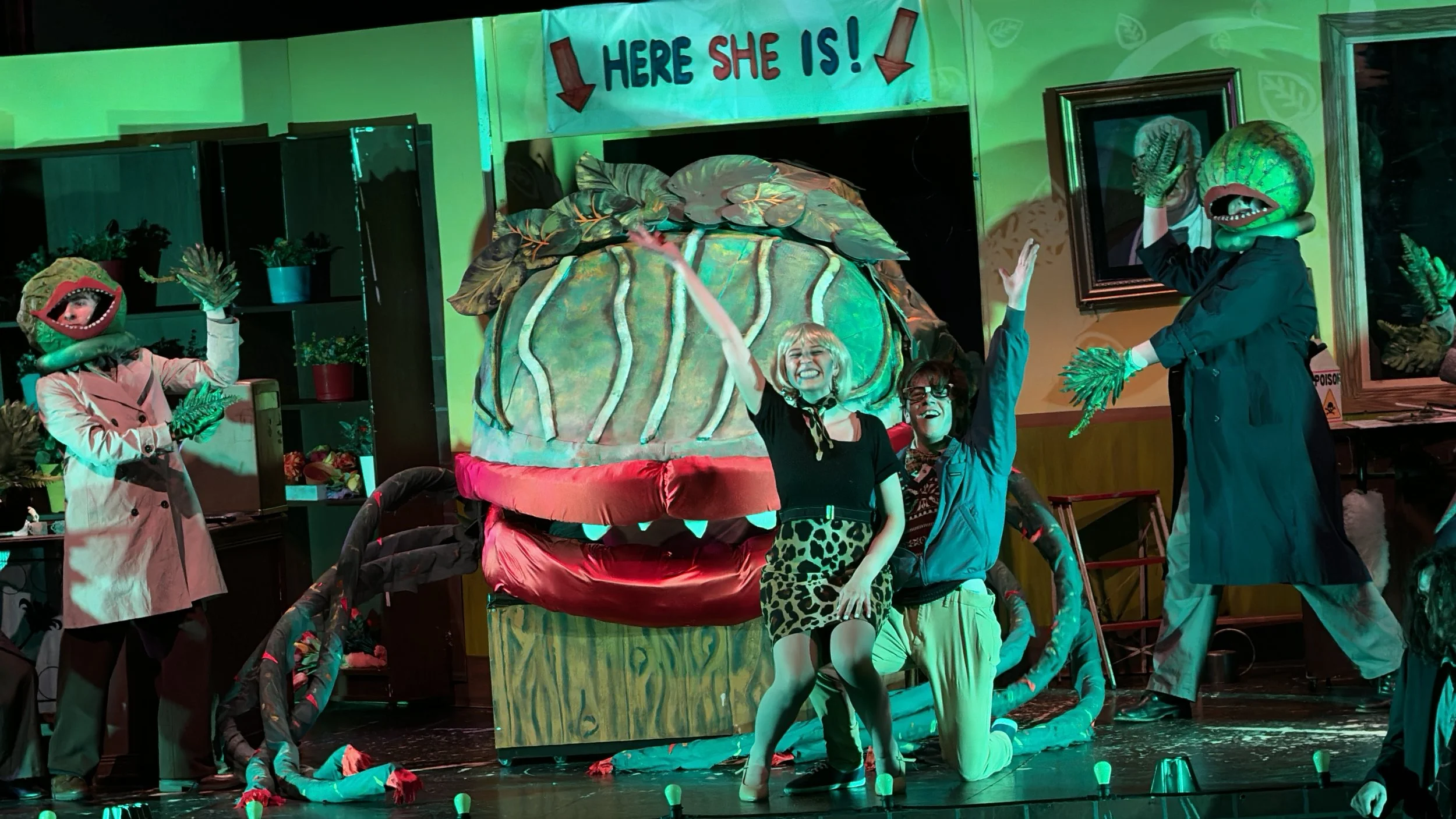 A theatrical stage scene with actors in colorful costumes, some with character masks or heads, performing around a large, whimsical Jack-in-the-Box prop. The background has a sign that says 'HERE SHE IS!' with arrows pointing down, and the stage is decorated with plants and framed pictures.