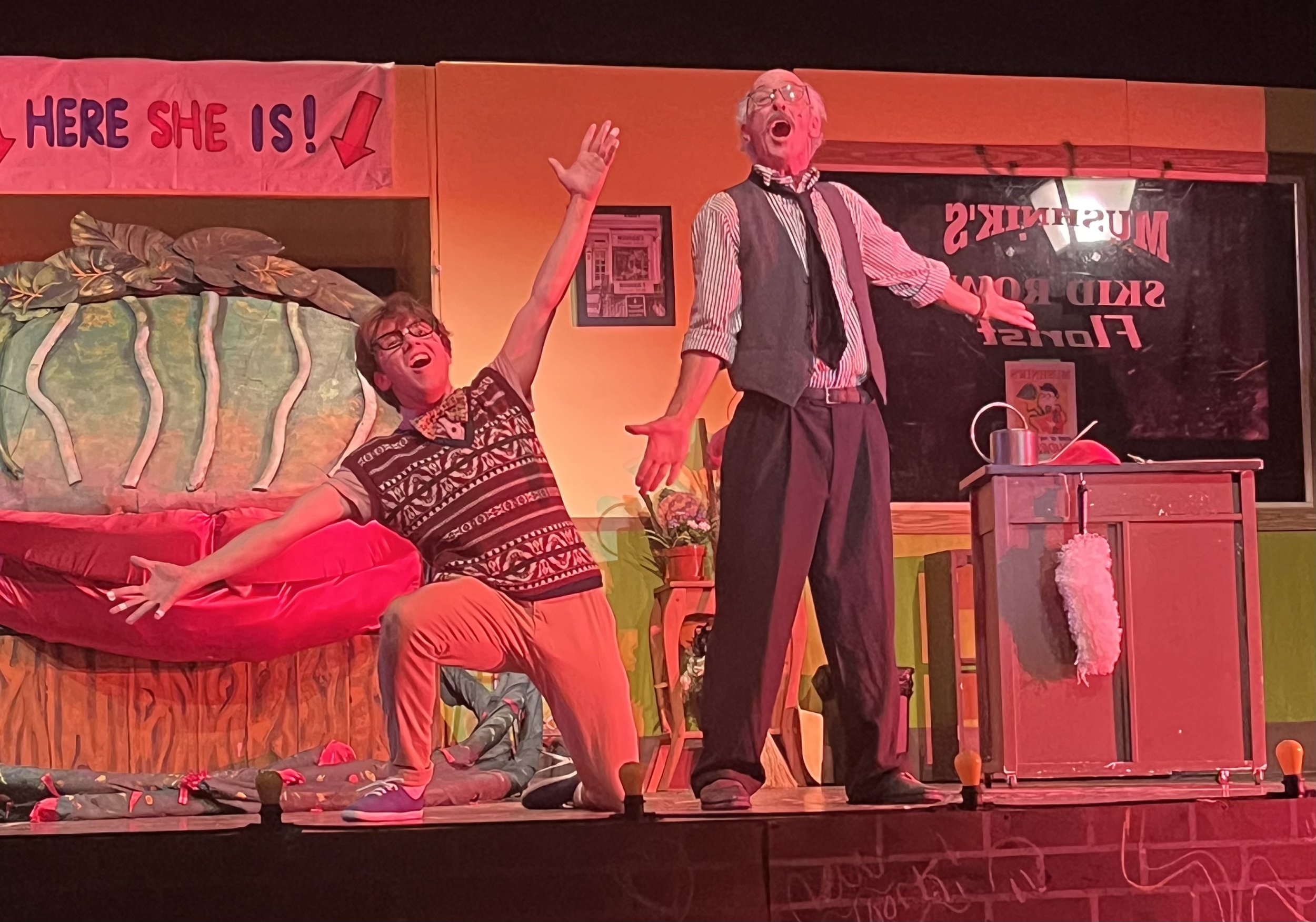 Two actors on stage performing in a play. One is kneeling with arms outstretched and face expressive, wearing glasses, a patterned shirt, and bright pants. The other, standing with arms open, also wearing glasses, a striped shirt, vest, and tie, is singing or speaking energetically. The stage has a colorful backdrop with a sign, a painted landscape, and props including plants and a cabinet.