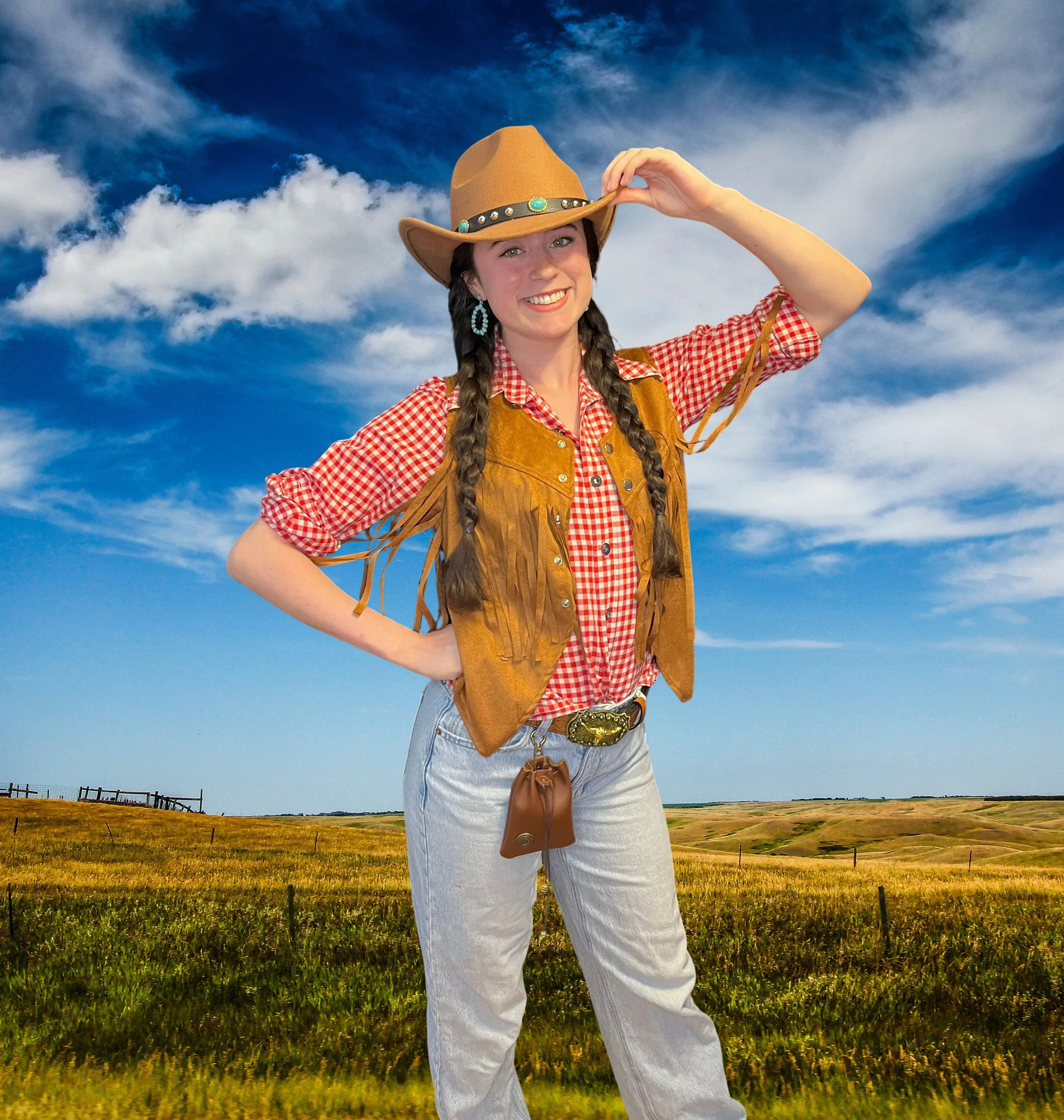 A young woman smiling outdoors in a grassy field under a cloudy sky. She is wearing a cowboy hat, turquoise earrings, a red checkered shirt, a brown fringed vest, and light denim jeans. She has long braided hair and is holding the brim of her hat with one hand.