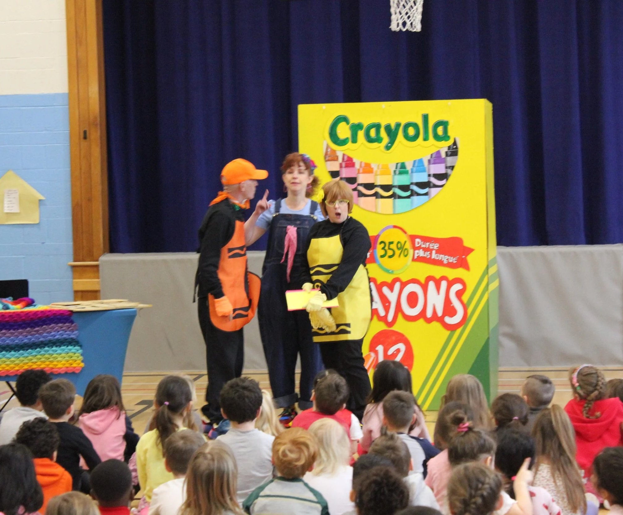 Three people dressed as Crayola crayons on stage with a large Crayola crayon box prop, speaking to children seated in the audience.