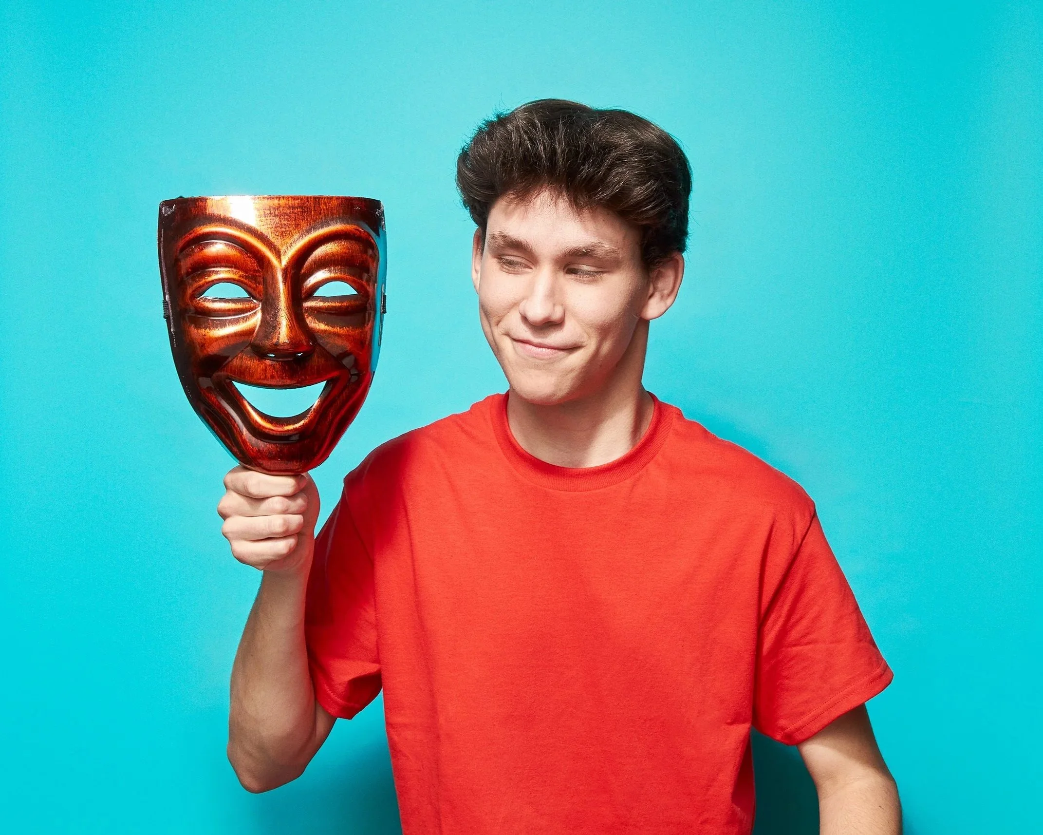 Man in red shirt holding a brown comedy mask against a blue background.