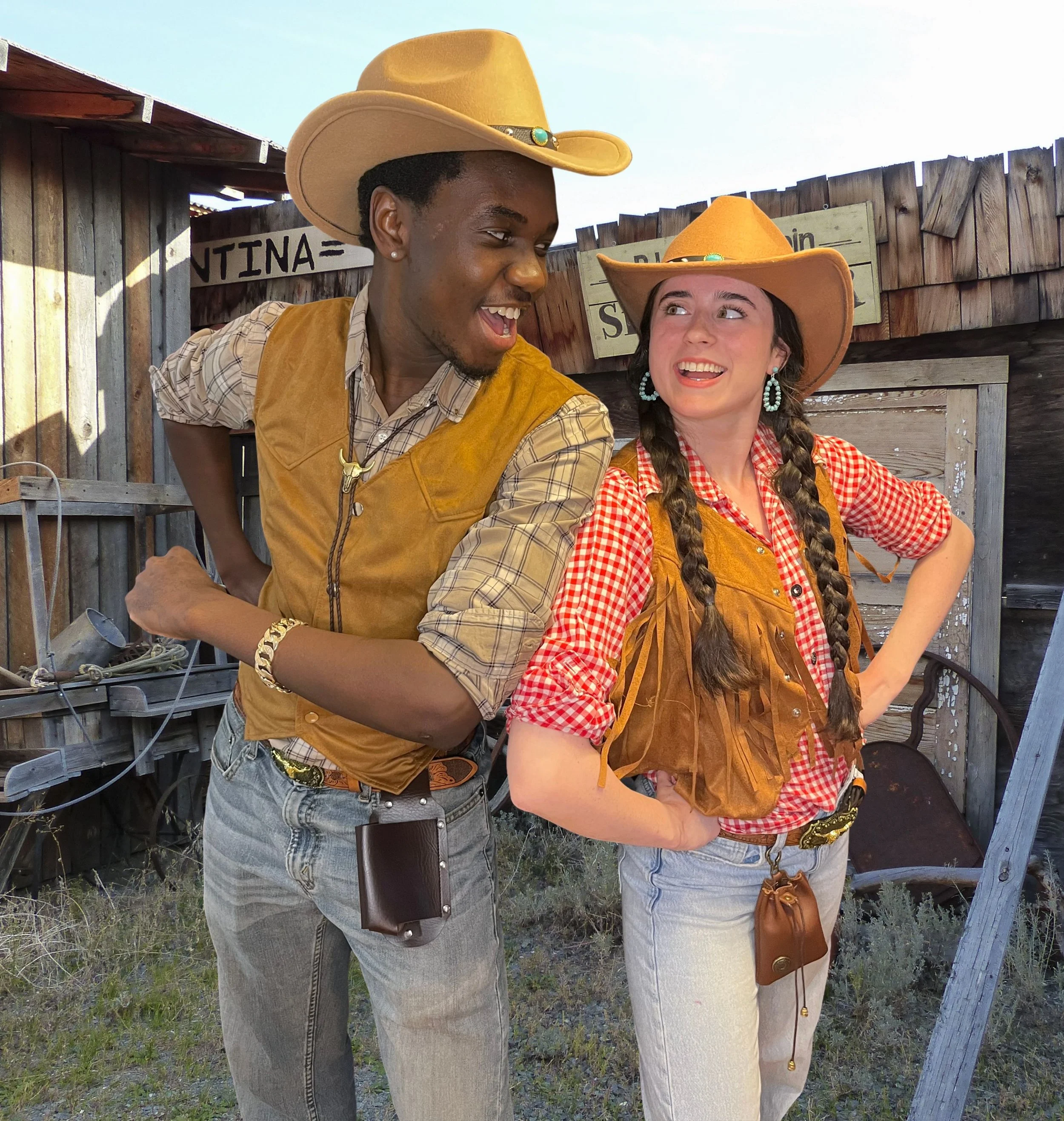 Two women in cowboy hats and western-style outfits standing outdoors near a wooden fence, smiling and looking at each other.