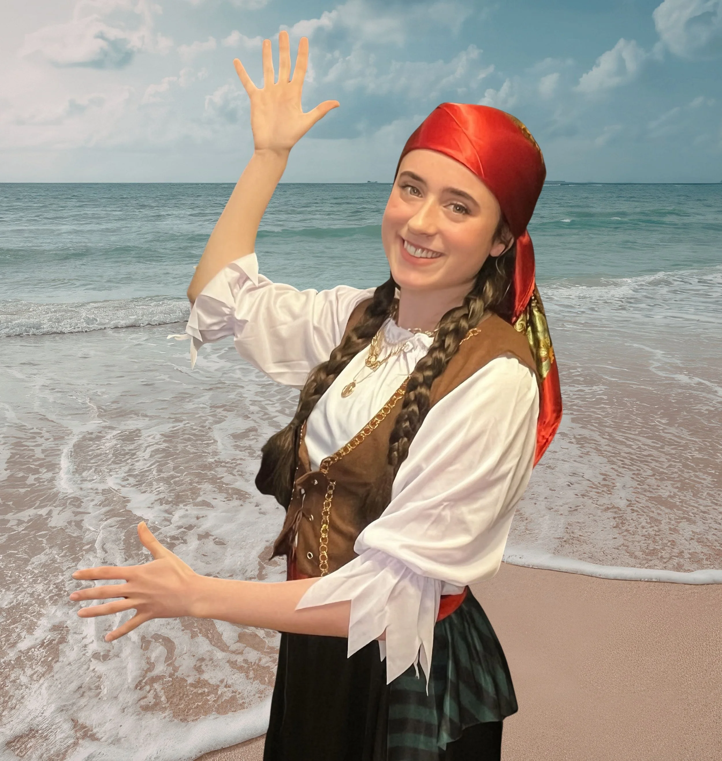 A woman in traditional pirate costume waving at the camera on a beach with ocean waves and a cloudy sky in the background.