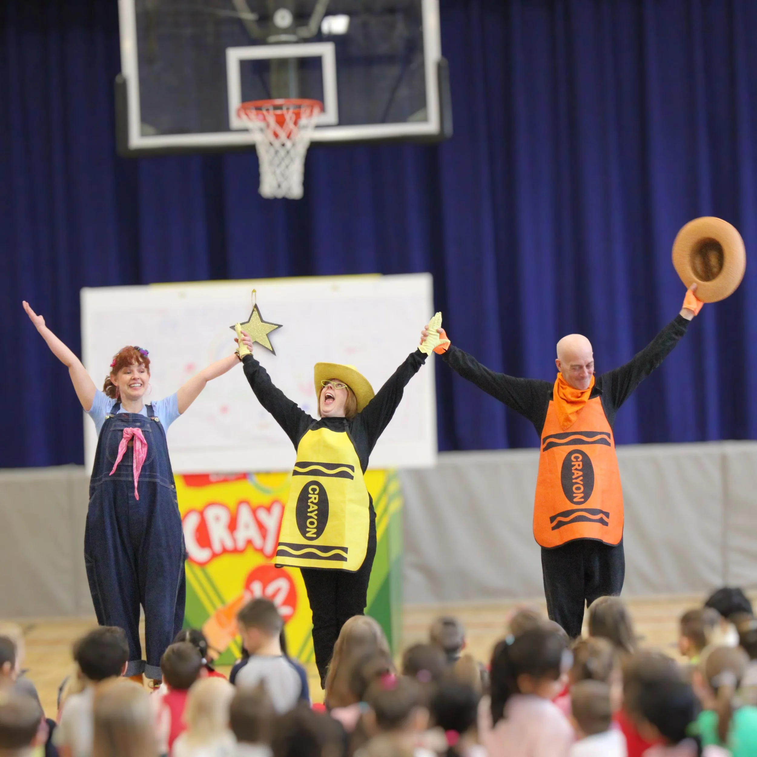 Three performers on stage entertaining children, one wearing a yellow Crayon costume, one in overalls , and the third in an orange Crayon costume. They are smiling and holding their arms up, with an audience of children seated on the floor.