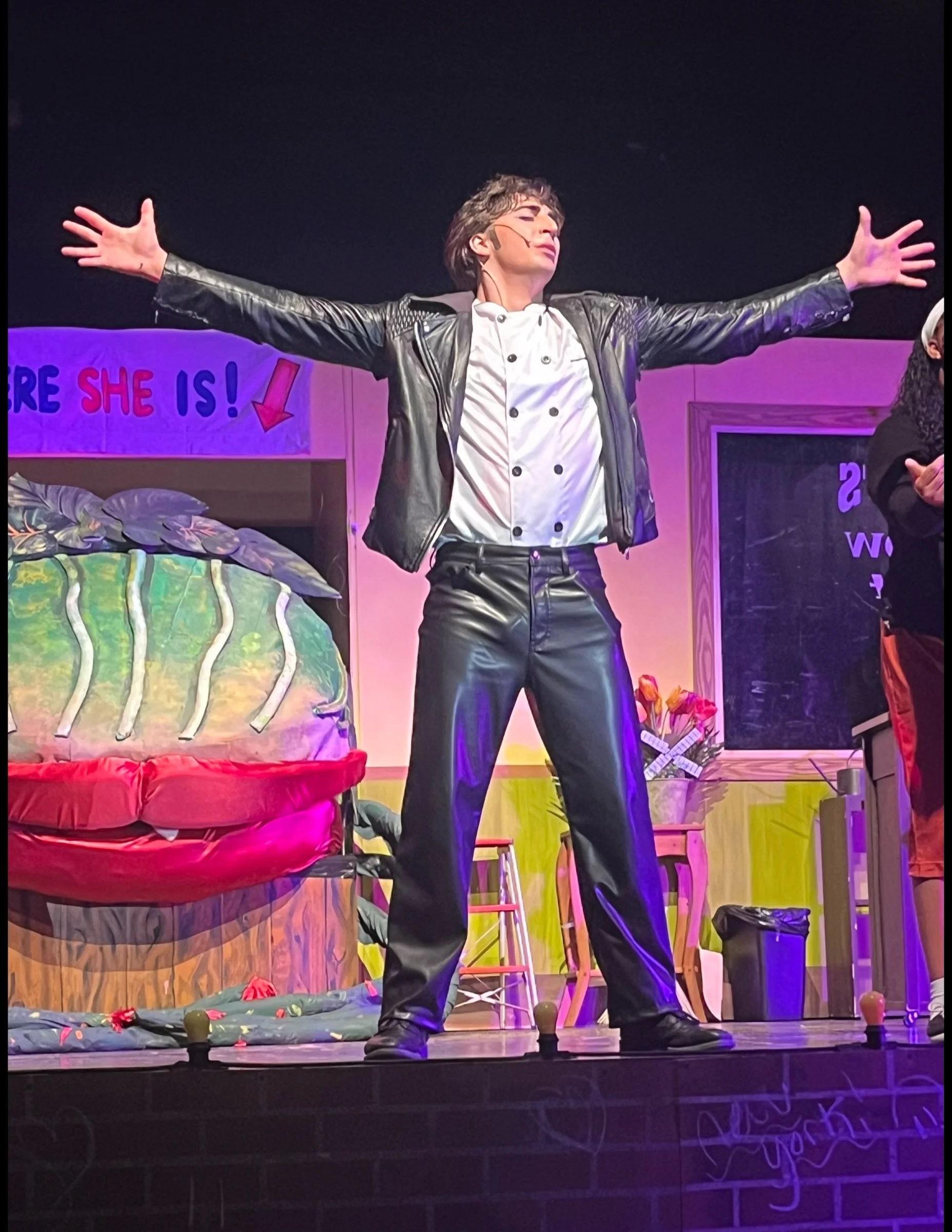 Performer on stage with arms outstretched, wearing a white shirt and black leather jacket and pants, in a theatrical setting with colorful props and a sign in the background.