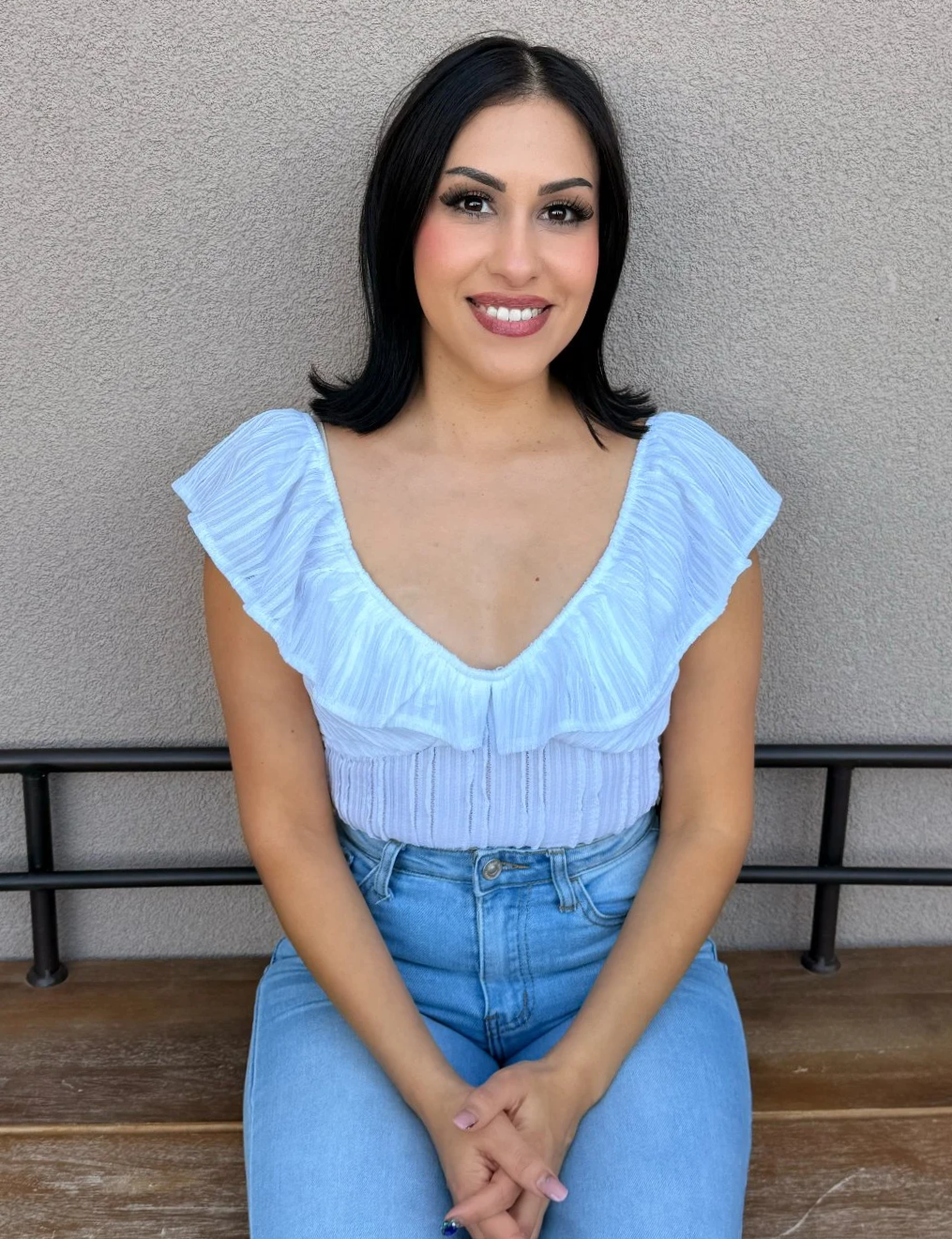 Woman with long black hair wearing an off-shoulder black top and white skirt, smiling in a light background.