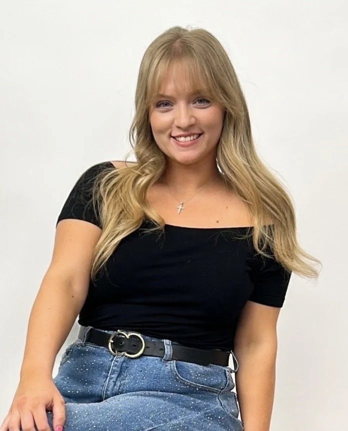 A young woman with long, wavy blonde hair, wearing a black t-shirt, blue jeans, a watch, and a necklace, standing in front of a blurred stone wall.
