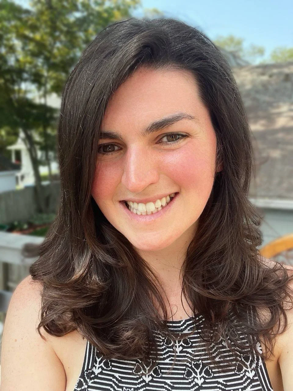 Smiling person outside with long dark hair and patterned sleeveless top, trees in the background.