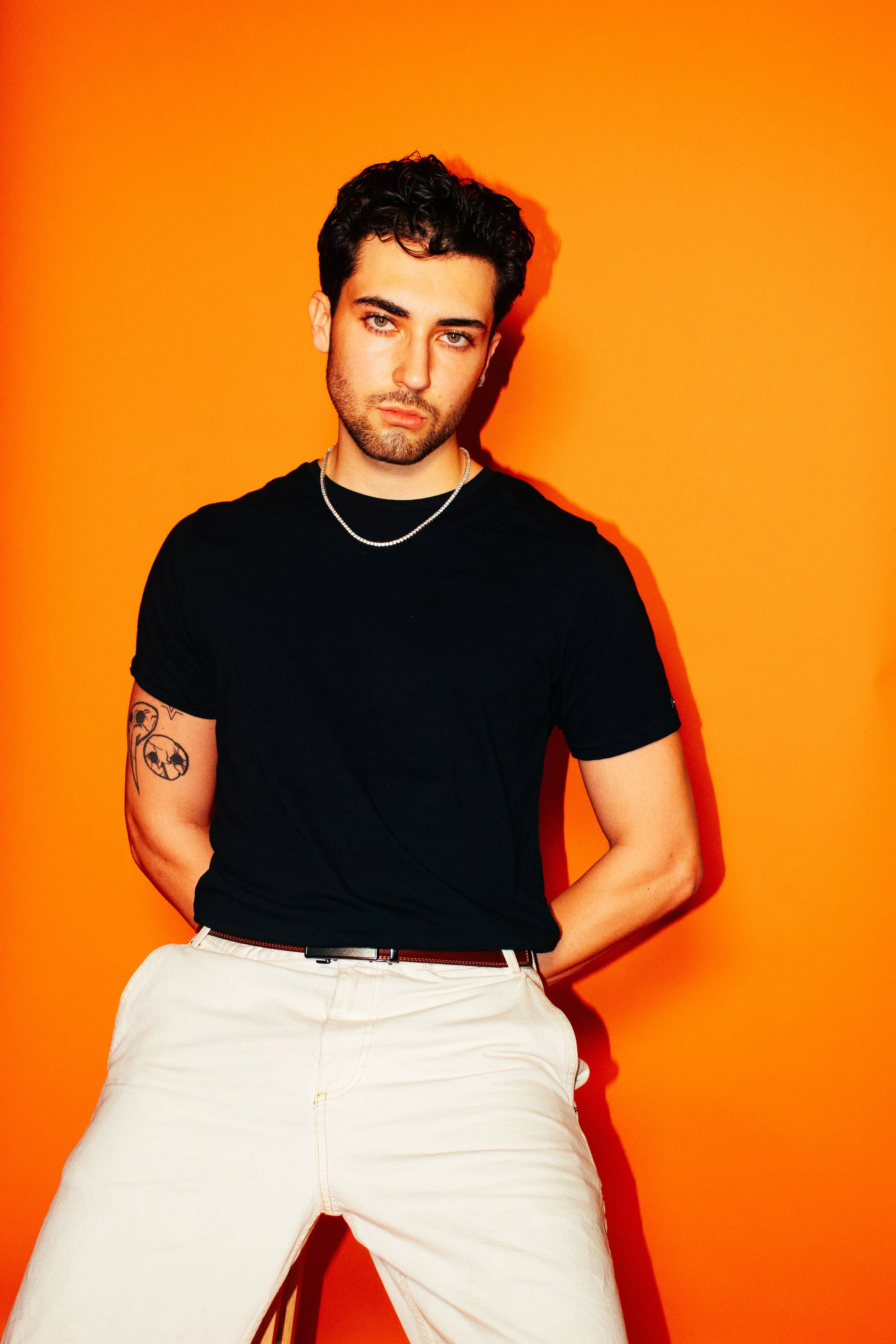 A young man with dark hair, wearing a black t-shirt, white pants, a silver chain necklace, and tattoos on his left arm, standing in front of a bright orange background.