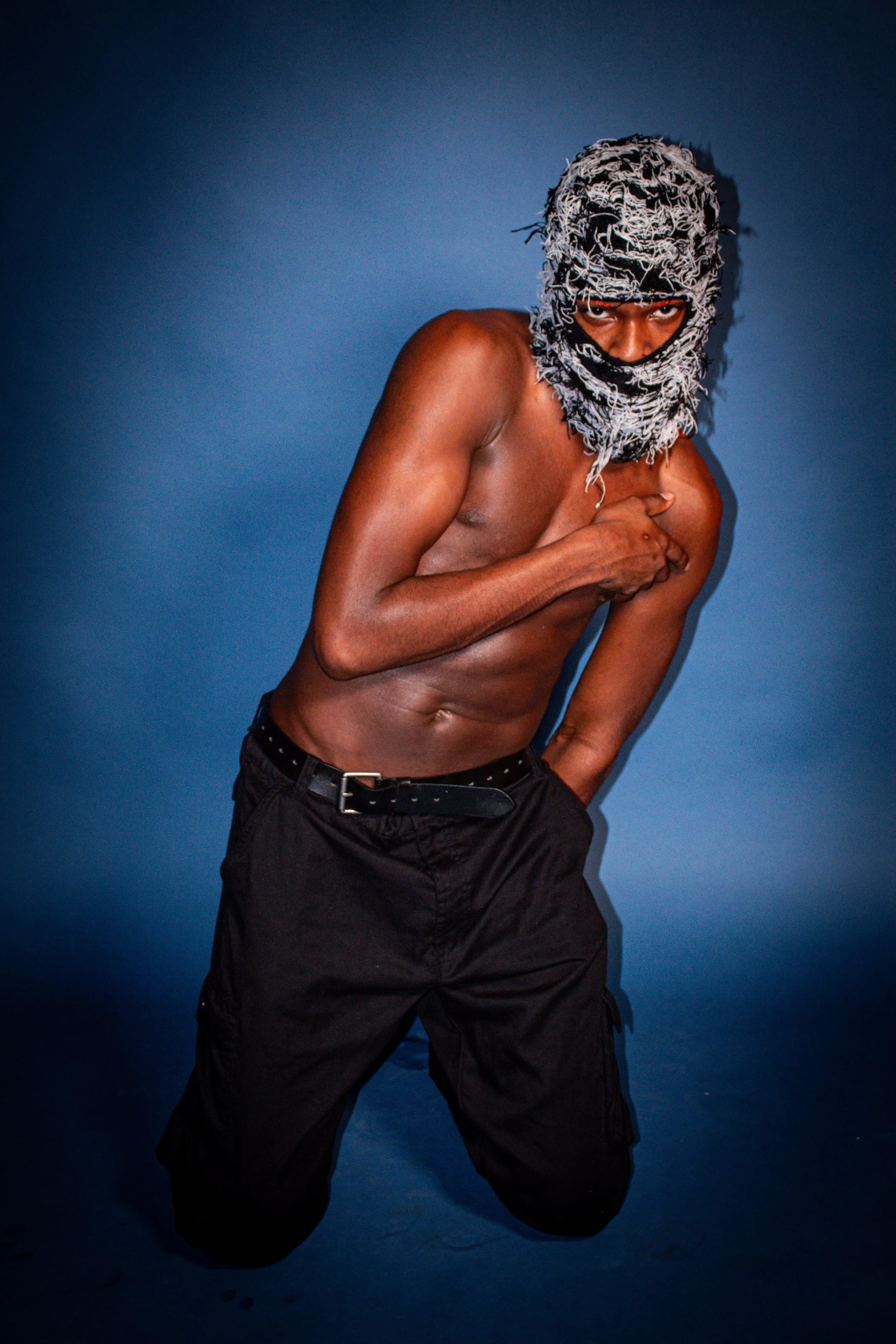 A shirtless man wearing a gray and white patterned mask covering his head and face, with only his eyes visible. He is posing against a dark blue background, with one hand touching his chest and the other resting on his waist, wearing black pants.