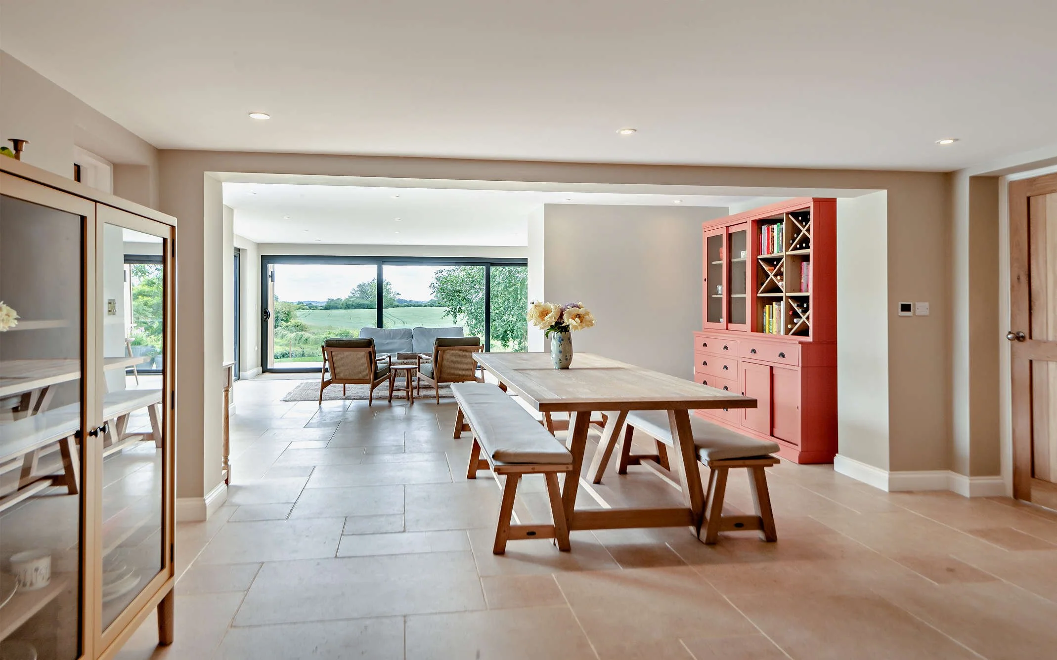 Open-concept living and dining area with wooden dining table, benches, a pink china cabinet, and large sliding glass doors leading to a green outdoor view.