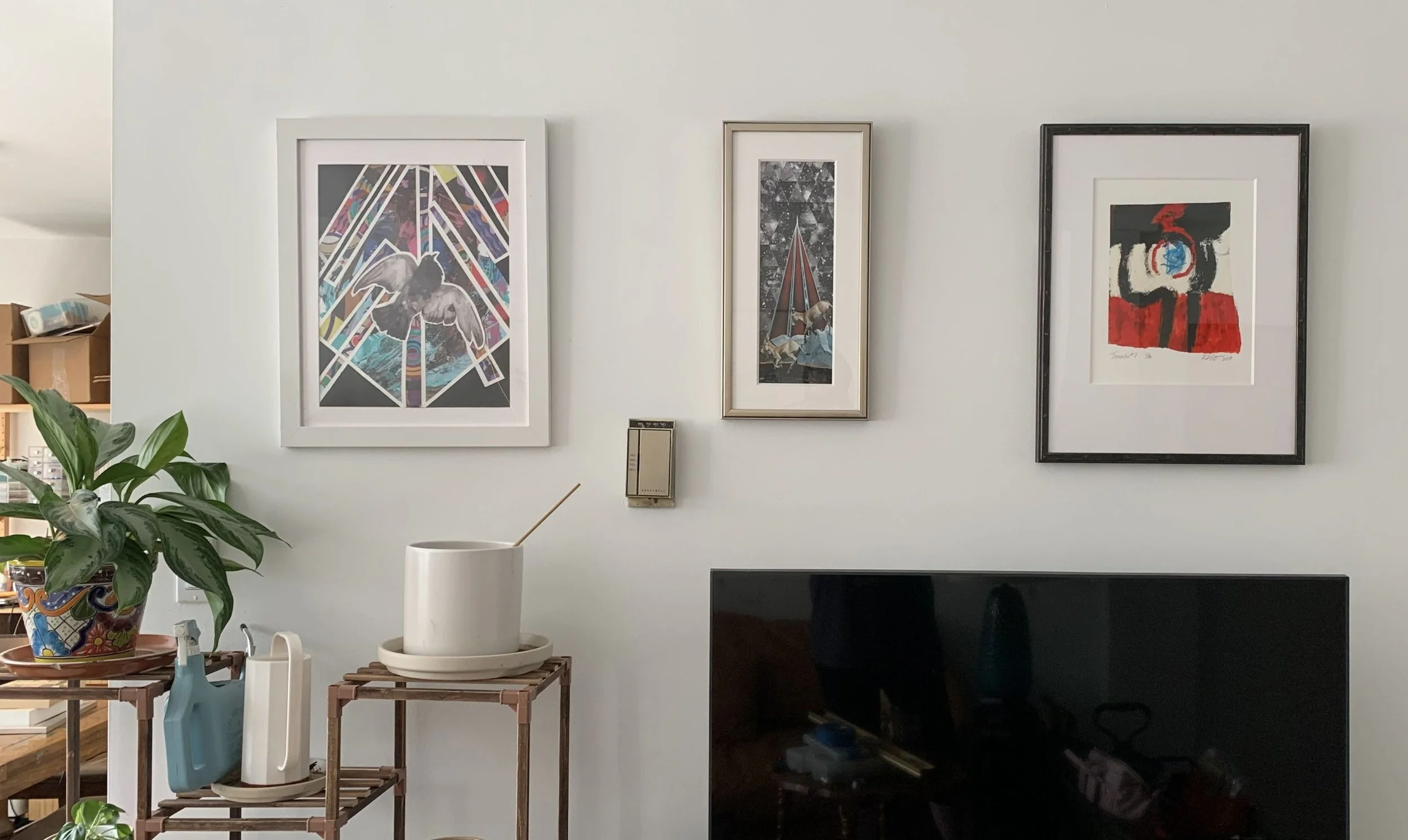 whie wall with 3 pieces of vertical framed artworks hung above a black fireplace and several plant stands. 