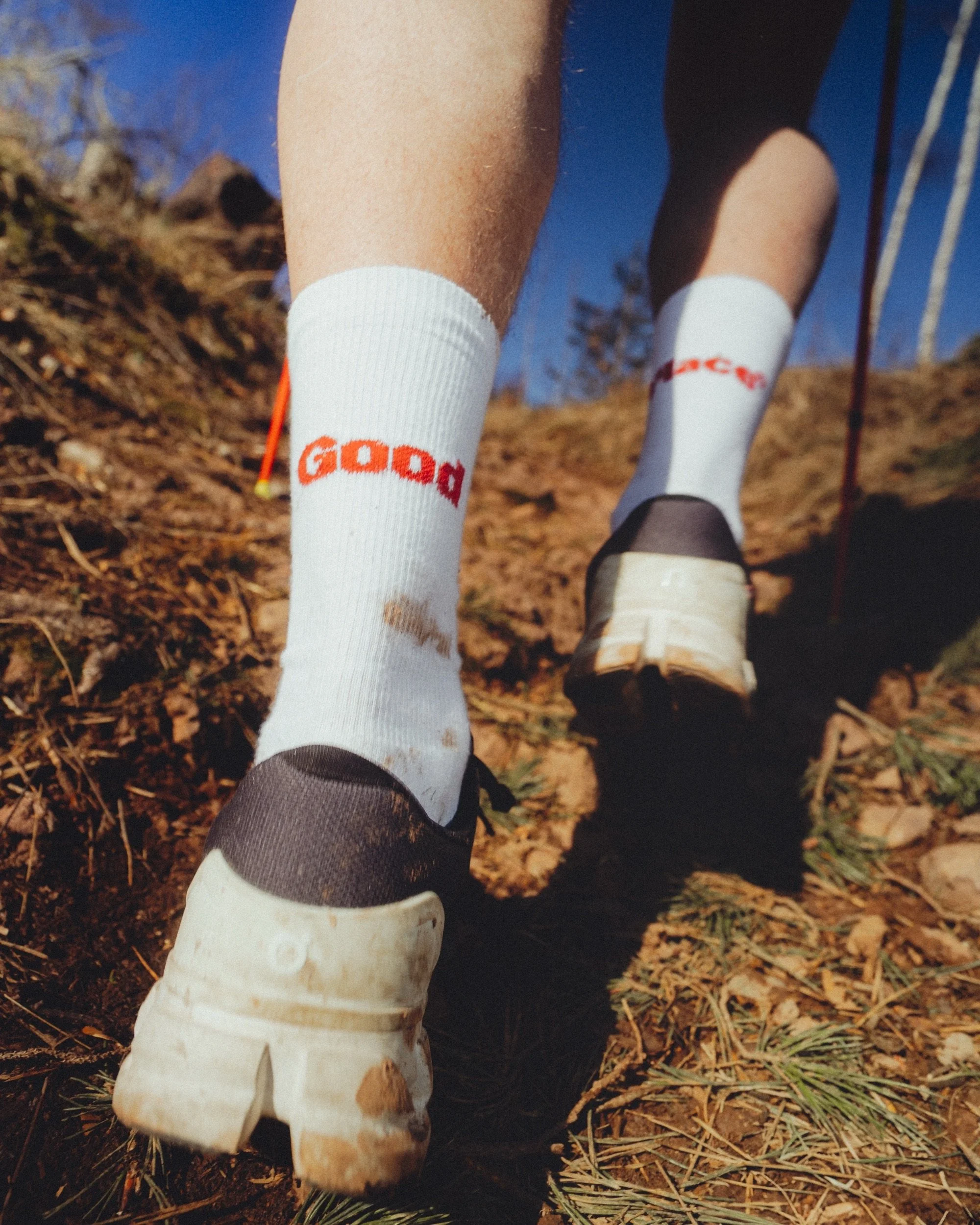 Close-up of a person's legs walking on a dirt trail with pine needles, wearing white socks with orange and red text and white sneakers, in an outdoor natural setting with trees and soil. Finishing in a good place socks close up trailrunning