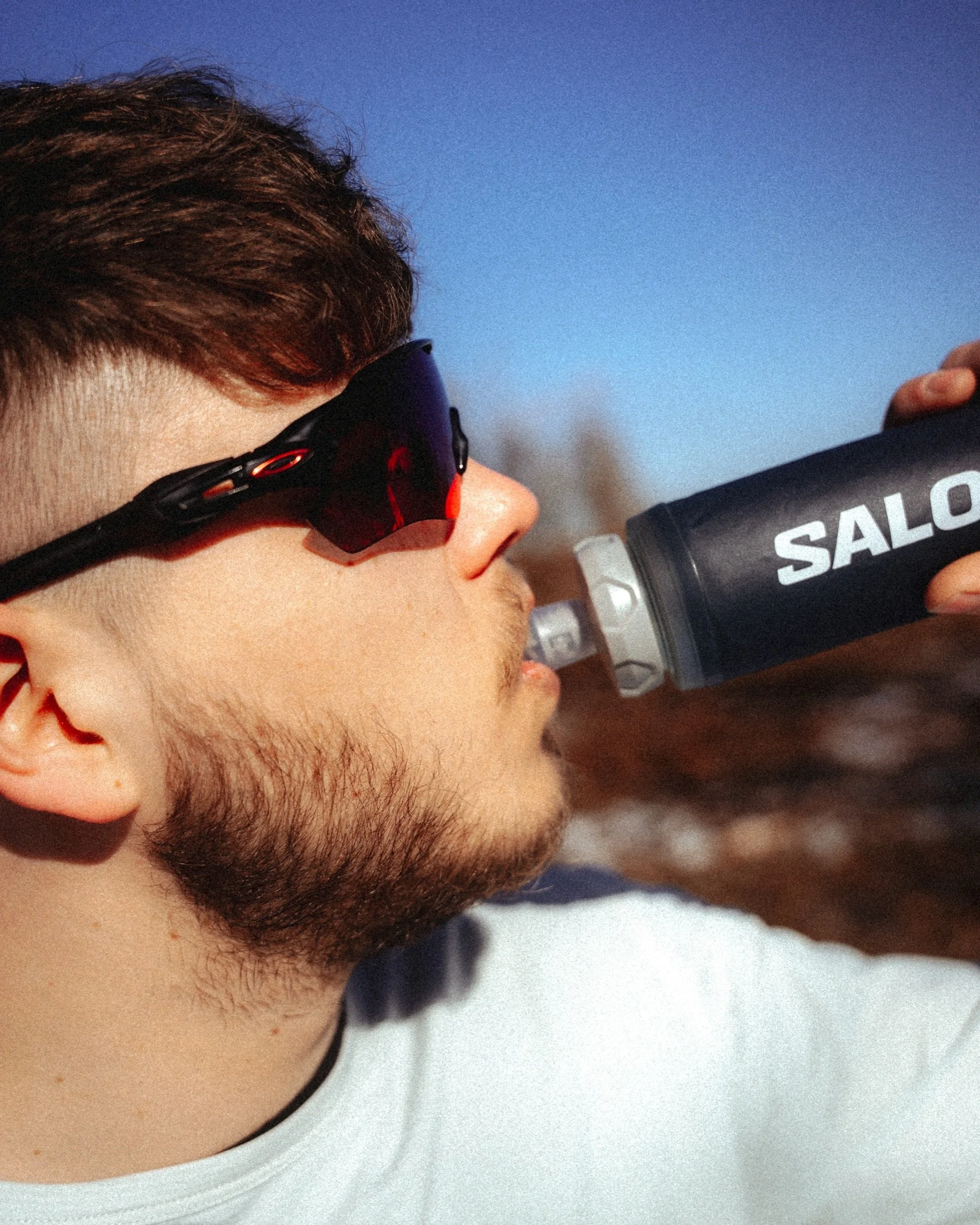 Close up photography from a trail runner in Oakley sunglasses drinking from a Salomon soft flask under a blue sky