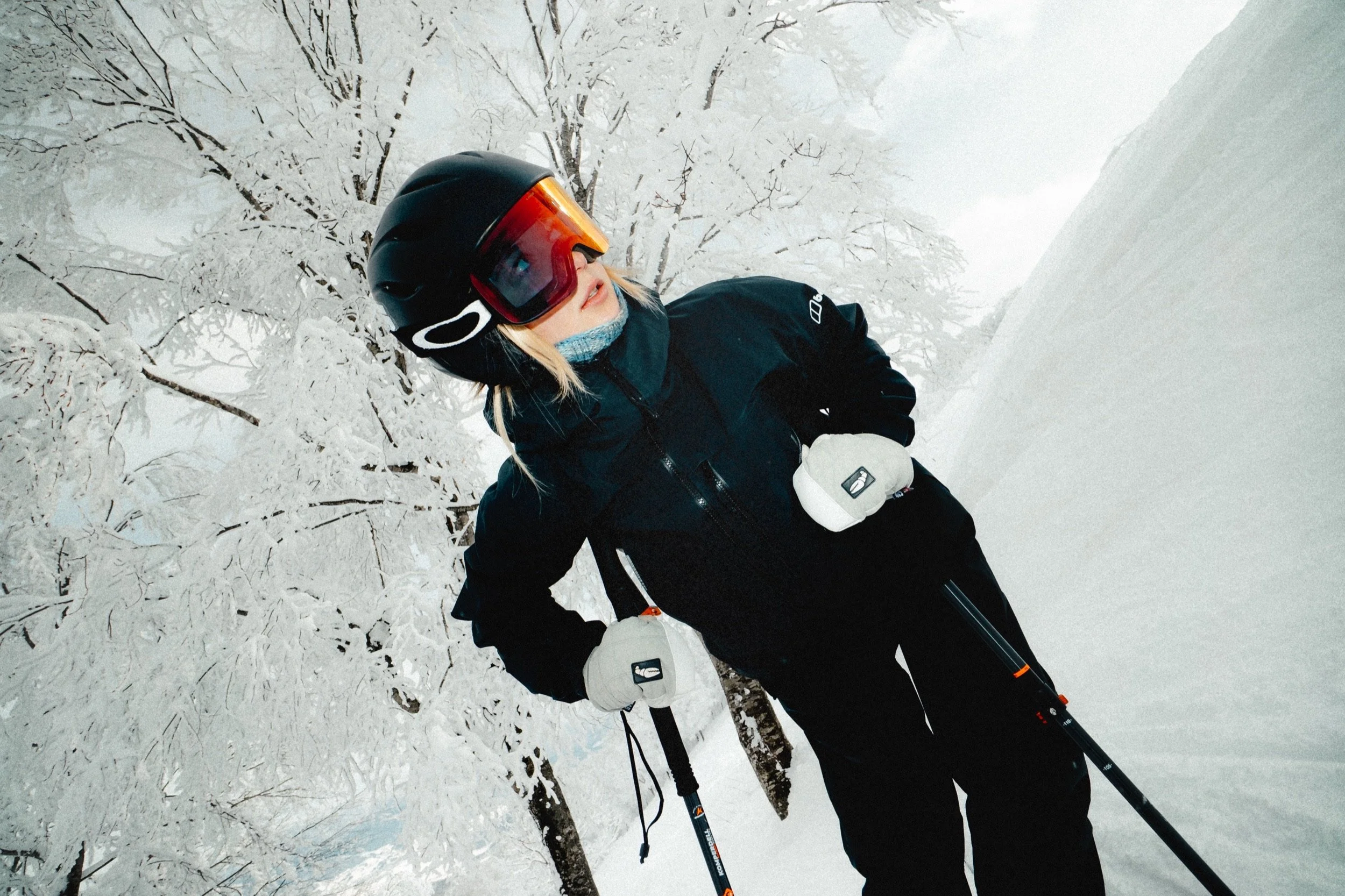 Person skiing in snowy forest, wearing a black helmet, ski goggles, black jacket, and white gloves, holding ski poles. Tangram, Madarao, Japan.
