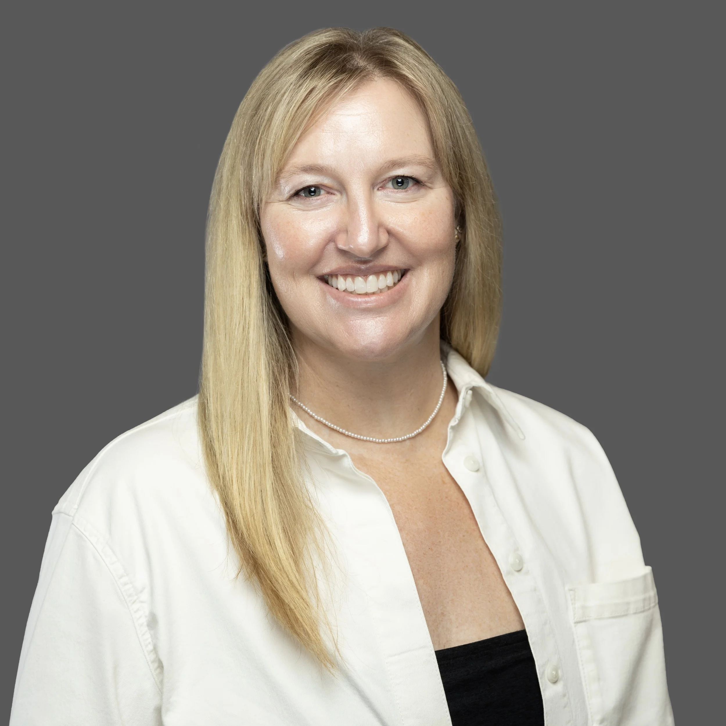 A smiling woman with long blonde hair, wearing a white button-up shirt, black top, and a pearl necklace, against a gray background.