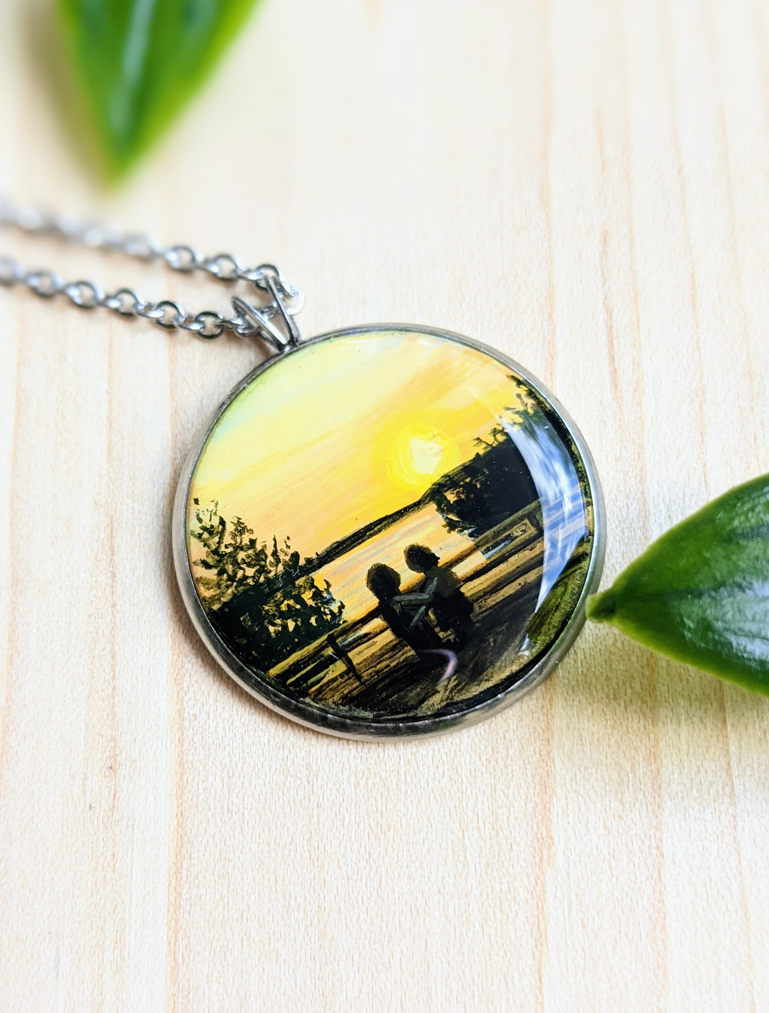 Custom hand-painted necklace featuring two silhouettes overlooking a lake at sunset (2022)