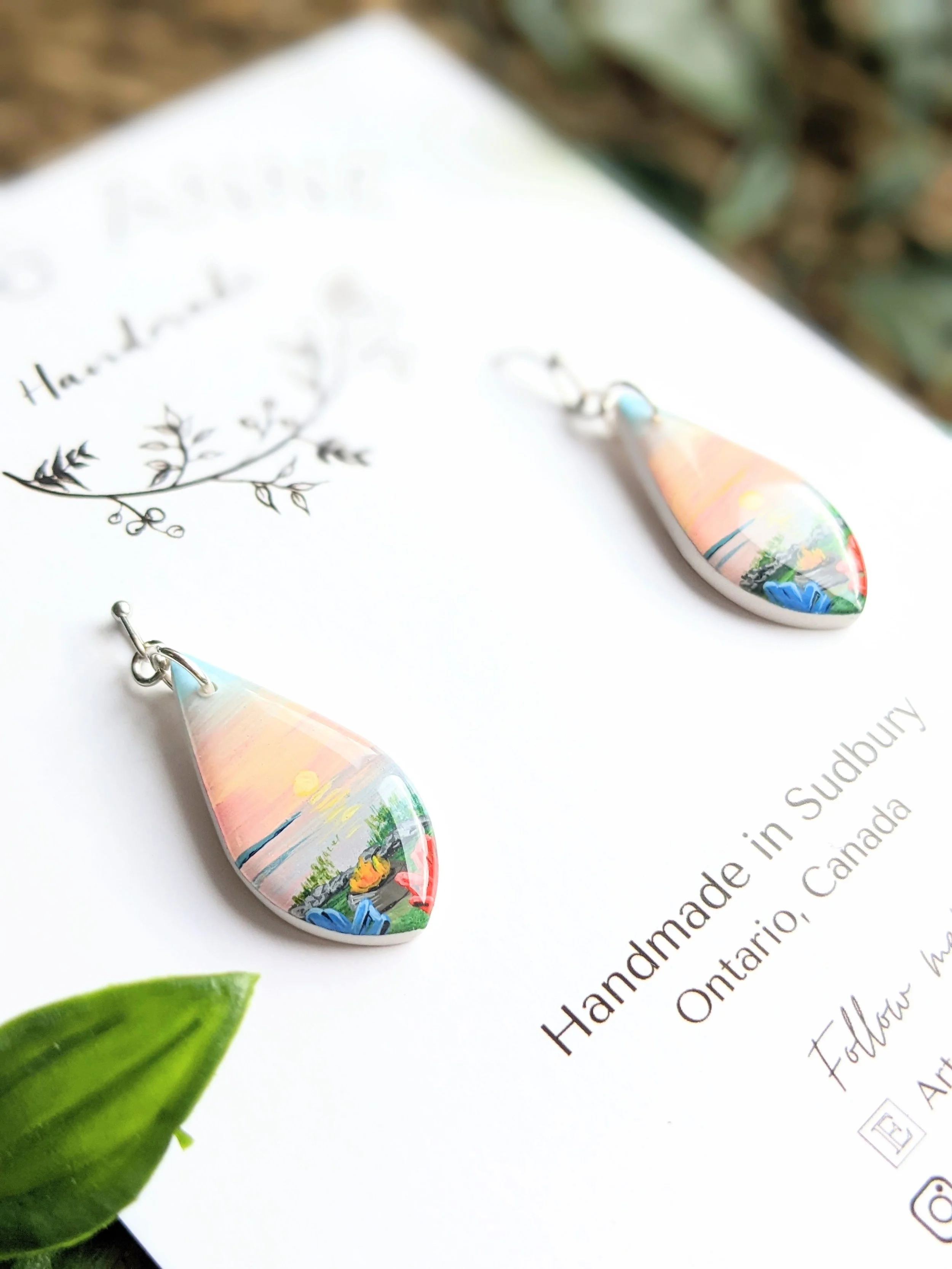 Custom hand-painted teardrop earrings featuring a lake at sunset, a campfire and two muskoka chairs facing the lake (2022)